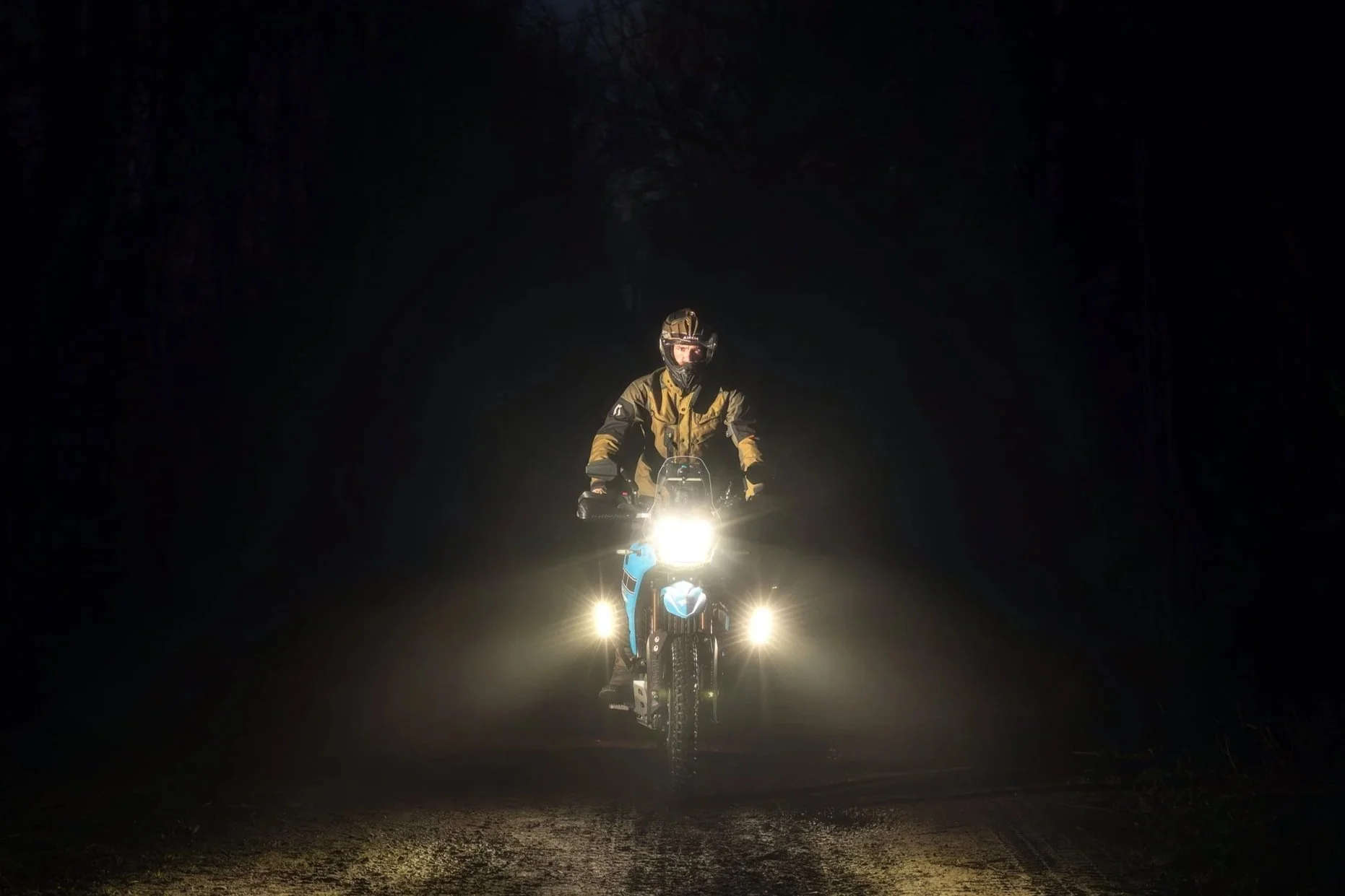 A person riding a motorcycle at night, illuminated by the motorcycle's headlights, with a dark forest background and a dirt road surface.