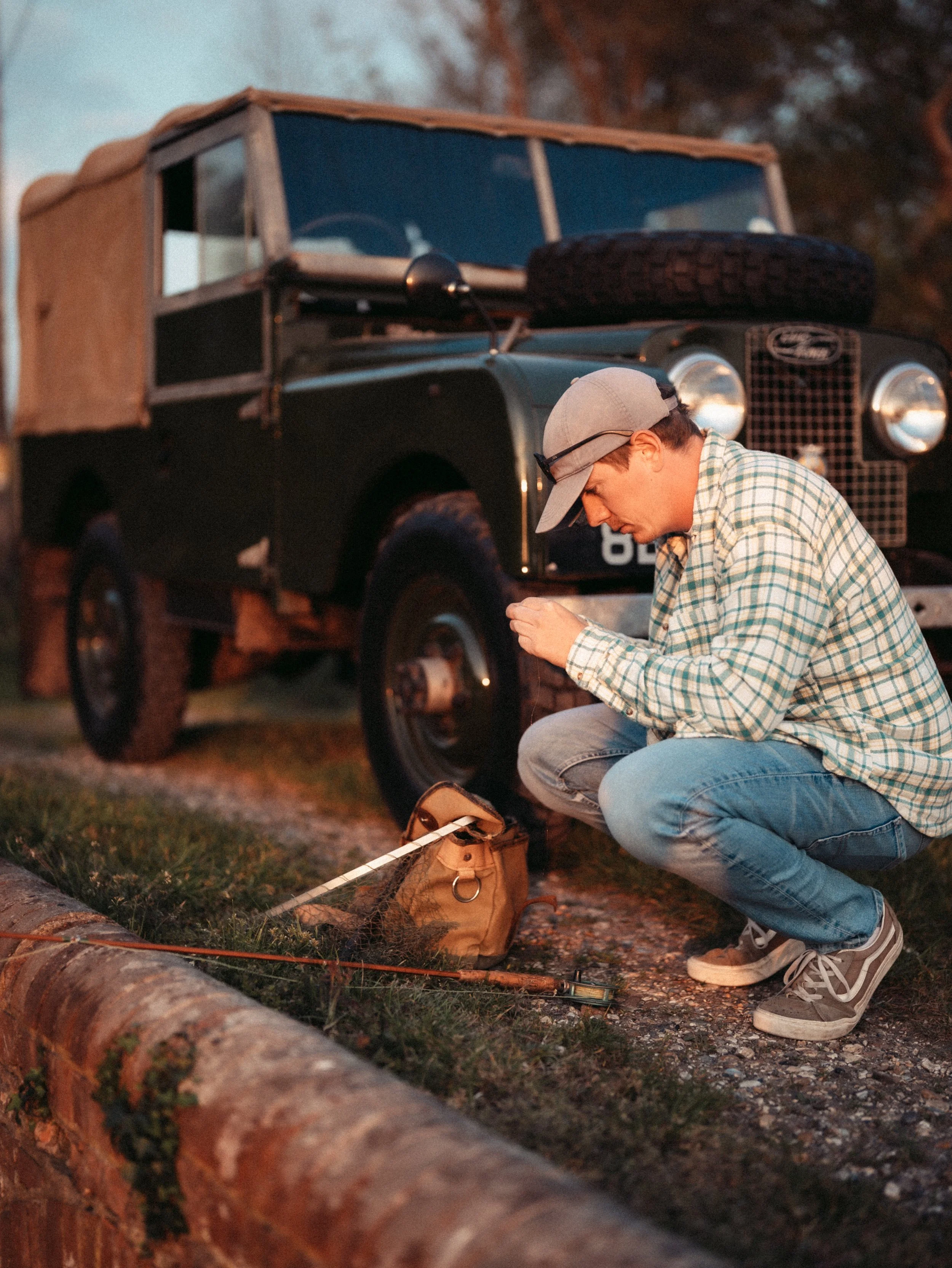 A young man wearing a plaid shirt, jeans, sneakers, and a baseball cap crouching on the ground, looking into a tan shoulder bag next to a fire hydrant. Behind him is a vintage Land Rover vehicle with a spare tire on the hood, parked on a grassy area 