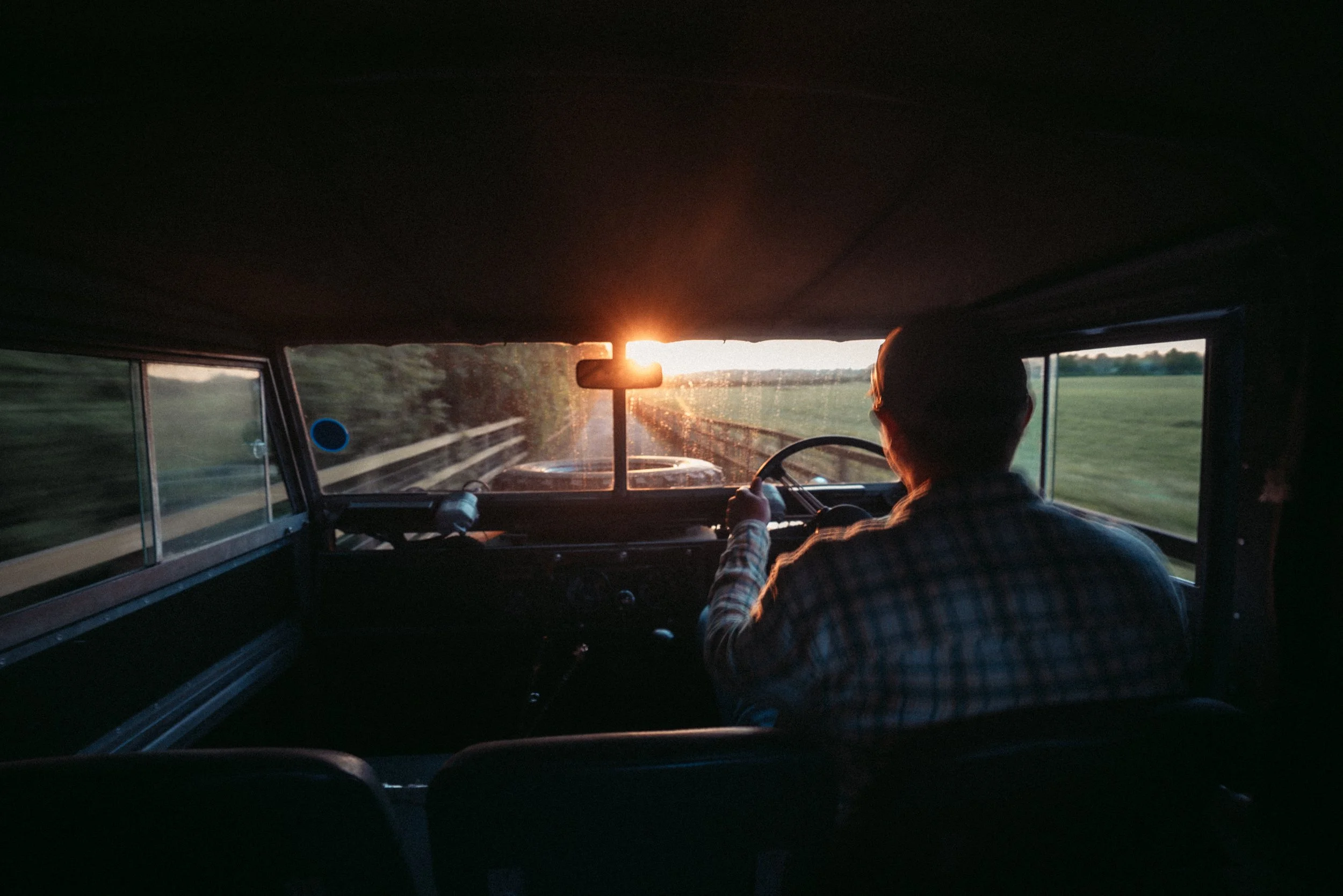 Inside a vehicle, two people are seen from behind, with the driver holding the steering wheel. The sun is setting on the horizon, casting a warm glow through the front windshield, illuminating a rural landscape with open fields and a fence.