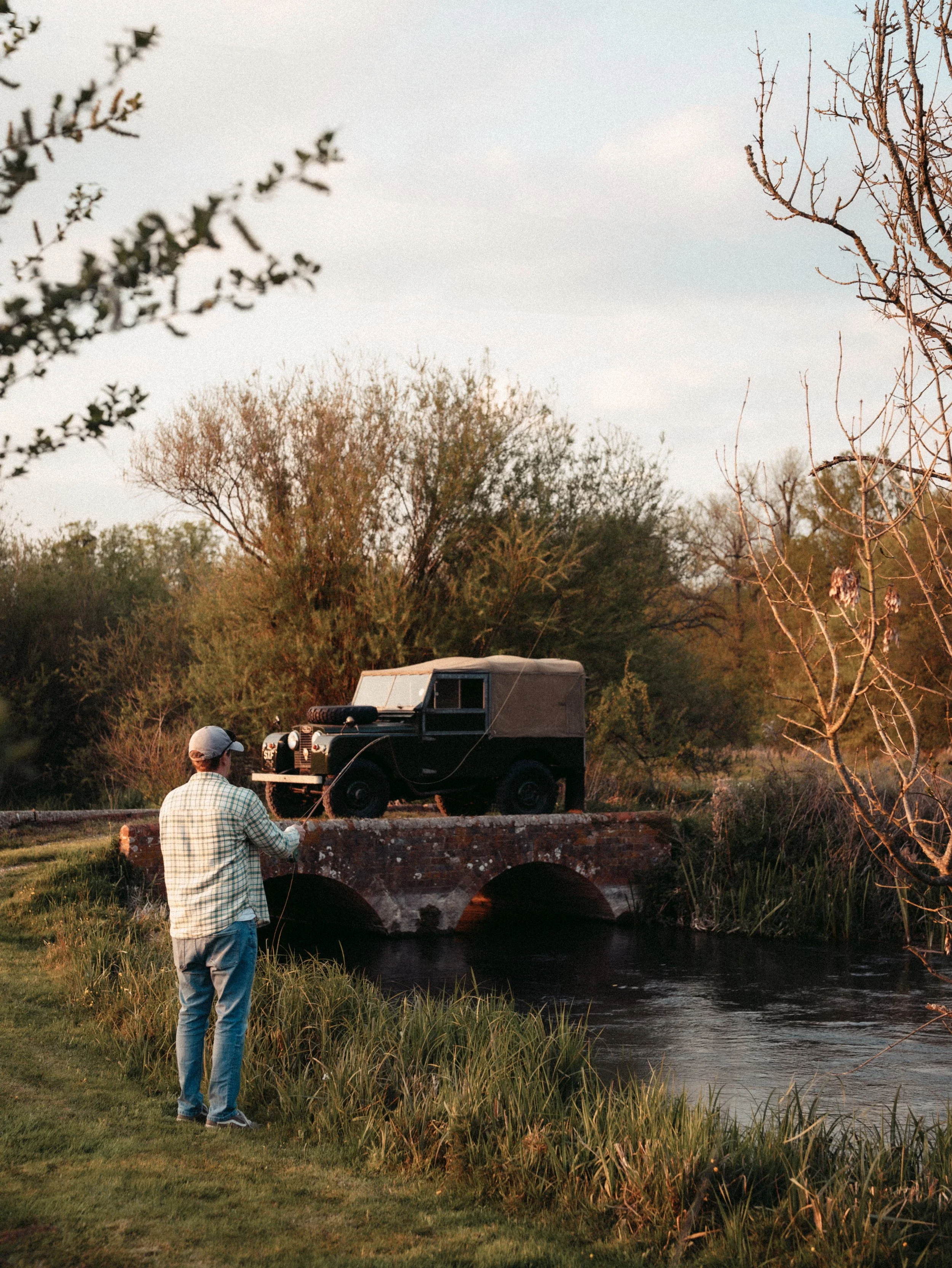 A man in a checkered shirt and baseball cap standing by a riverbank, fishing, with a vintage black truck parked on a brick bridge in the background, surrounded by trees and bushes.
