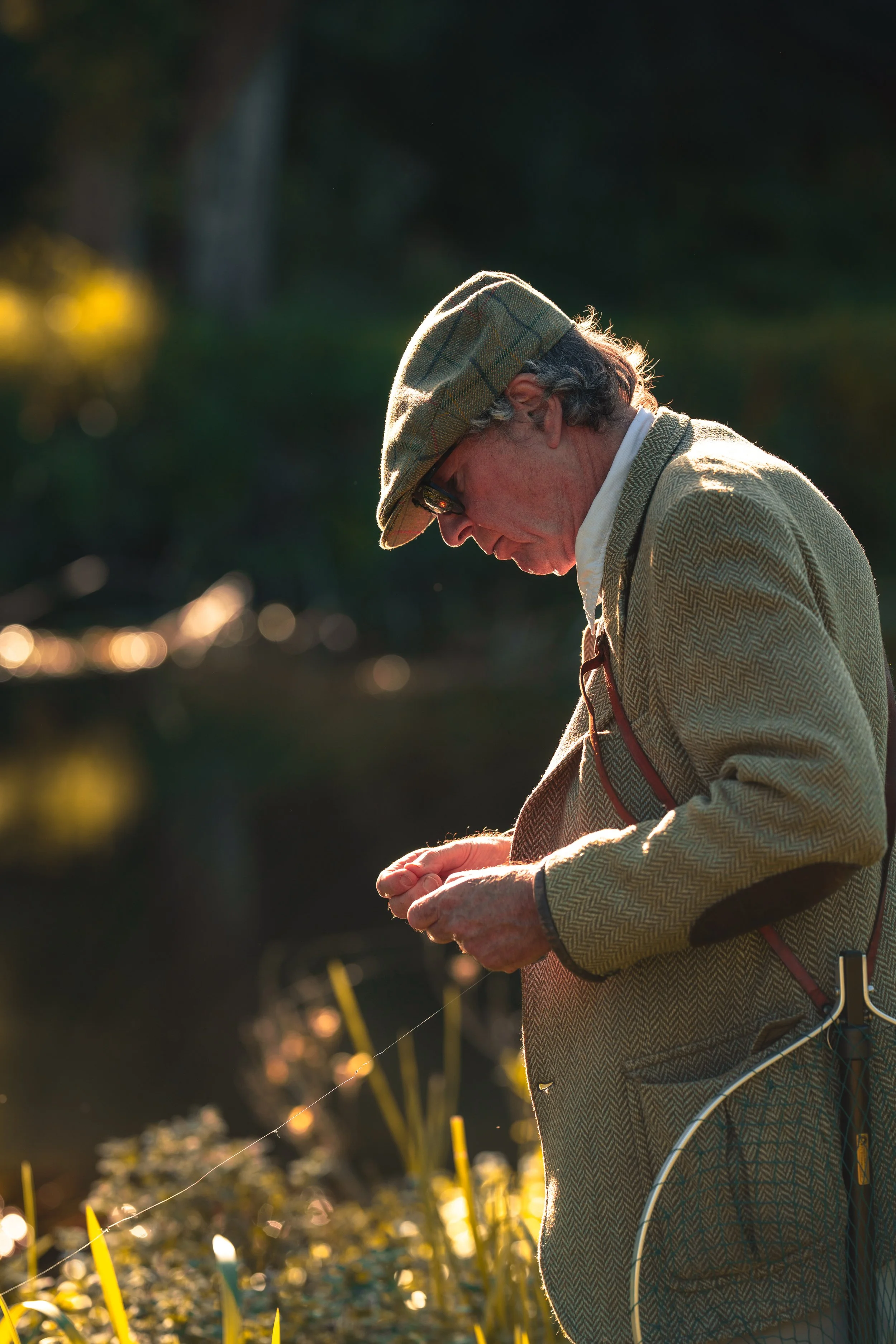 An elderly man wearing a plaid hat, glasses, and a beige jacket, is fishing in a natural setting during golden hour with sunlight creating a warm glow.