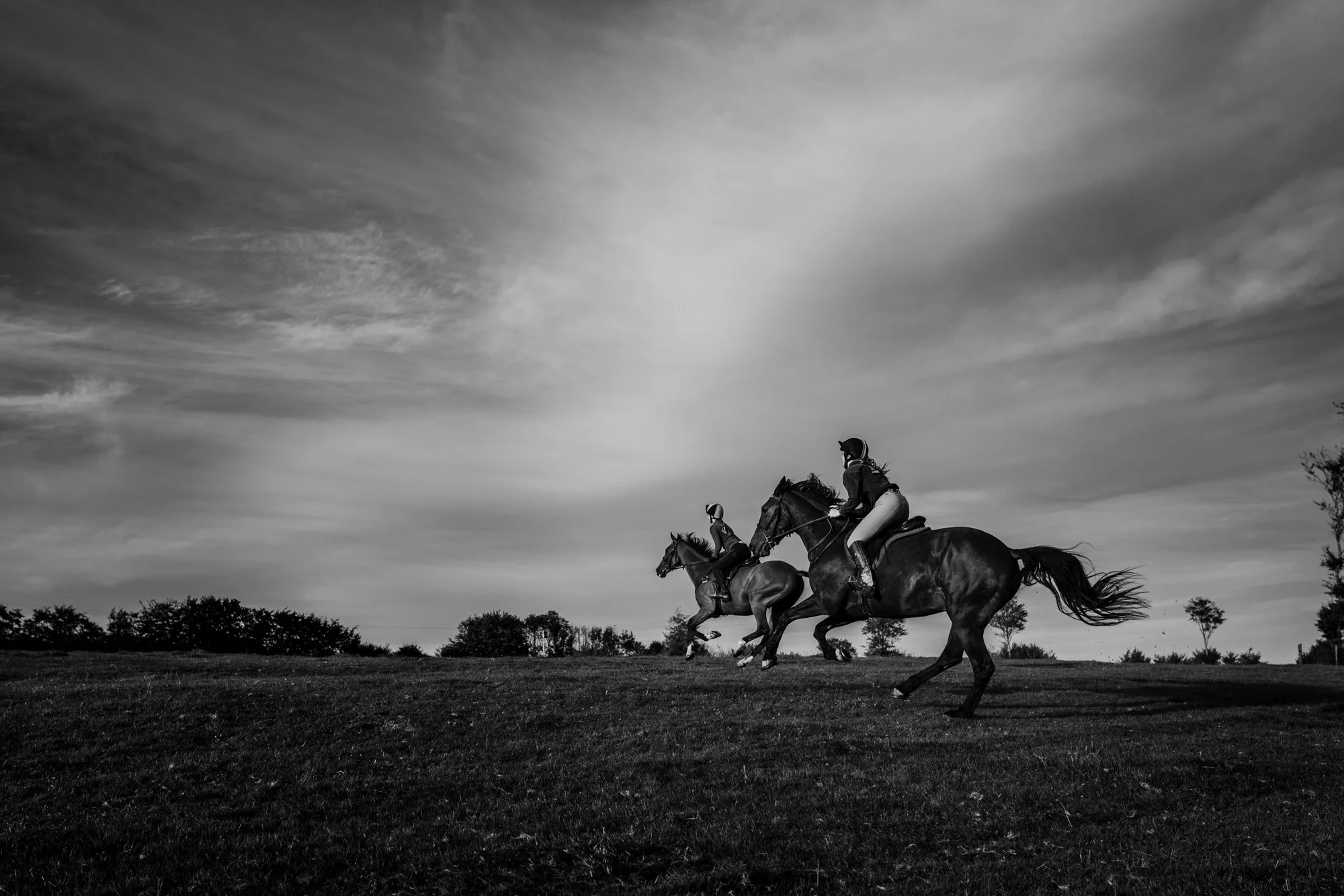 Black and white photo of three horseback riders riding across an open field under a sky with clouds.