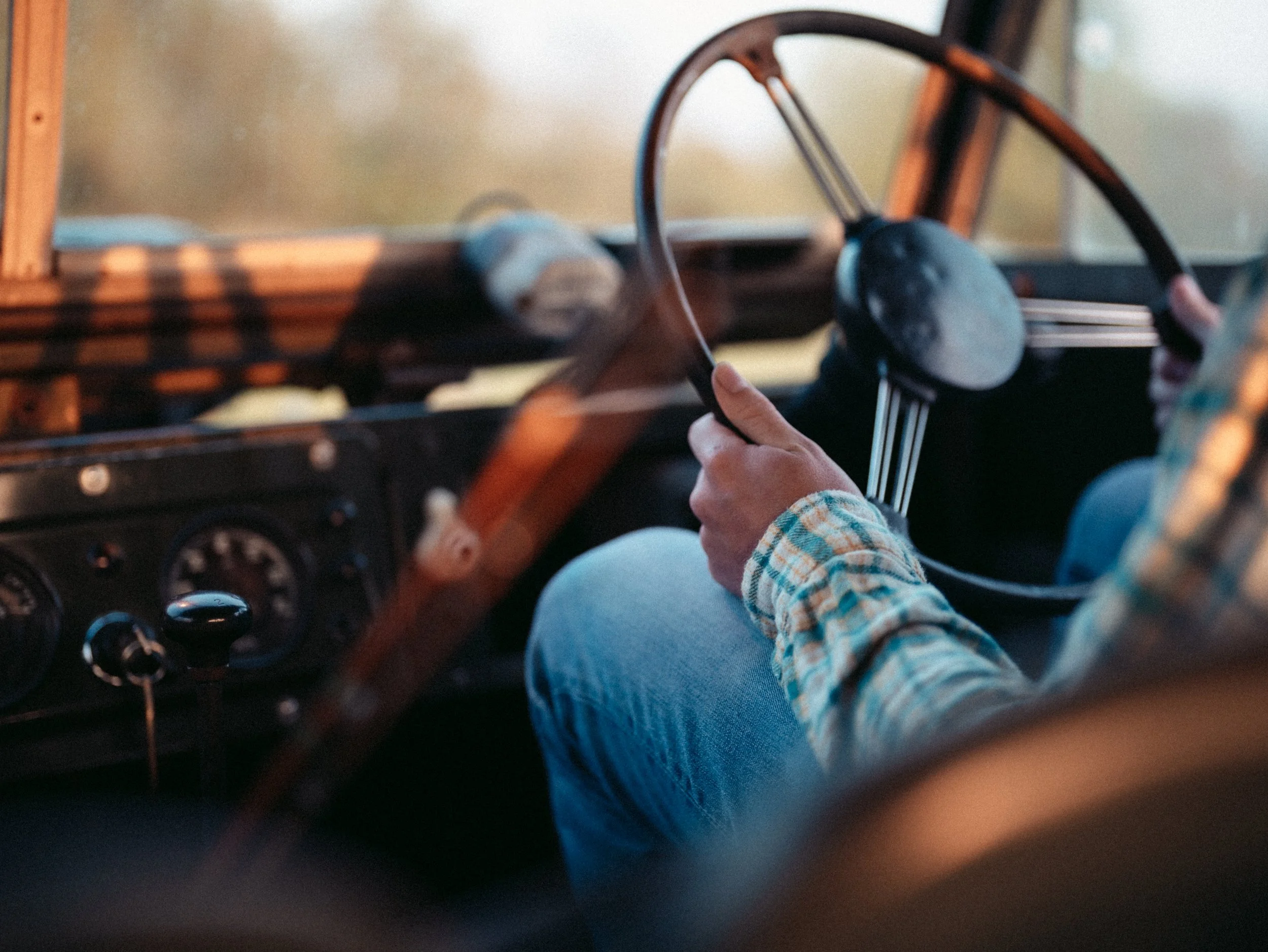 Close-up of a person wearing a checkered shirt and blue jeans, holding a steering wheel in a vintage vehicle, with blurred dashboard and windows in the background.