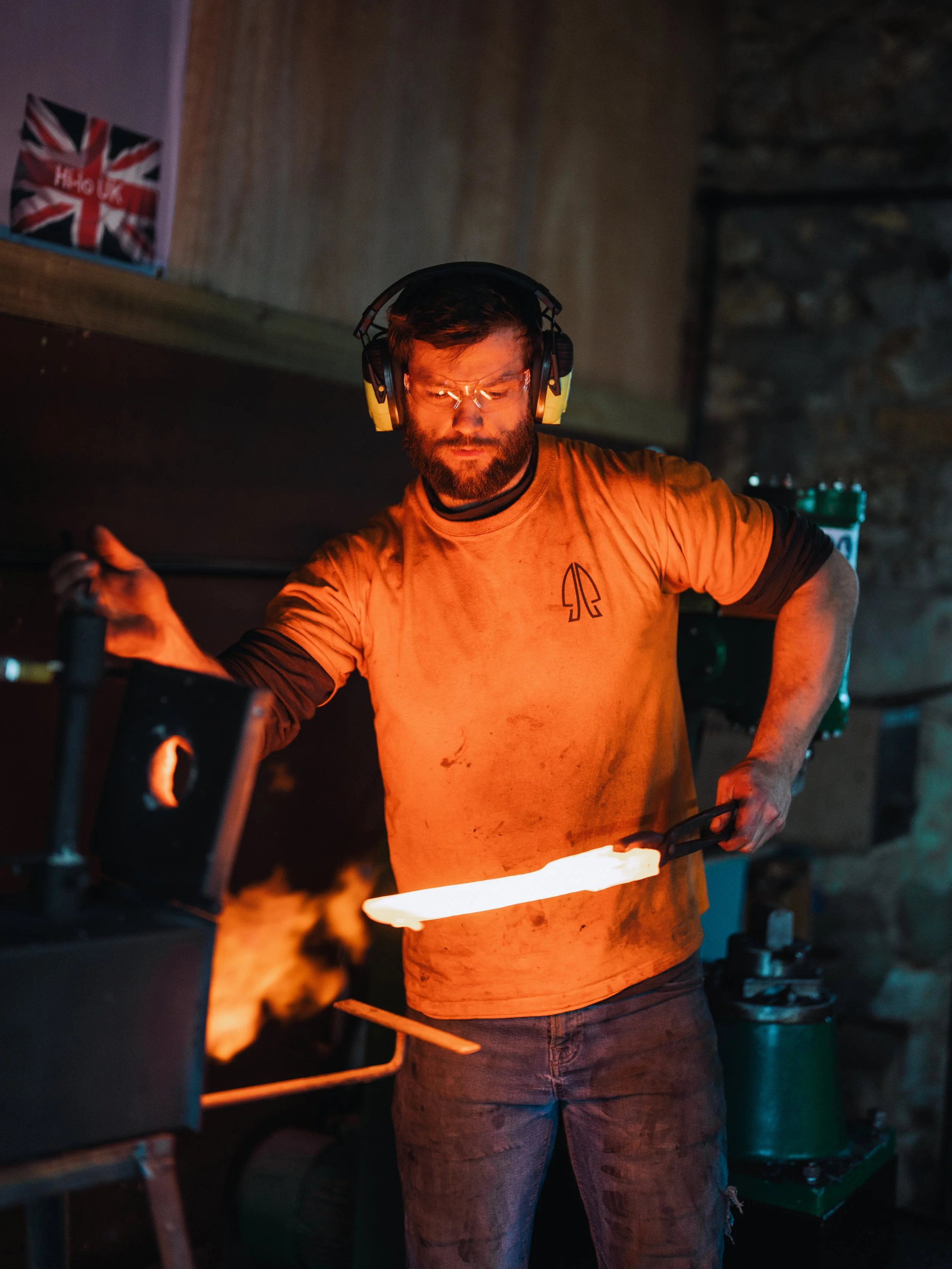 A man wearing safety glasses and ear protection is forging a hot metal piece in a blacksmith workshop, with a background that includes a stone wall and a poster of the British flag.