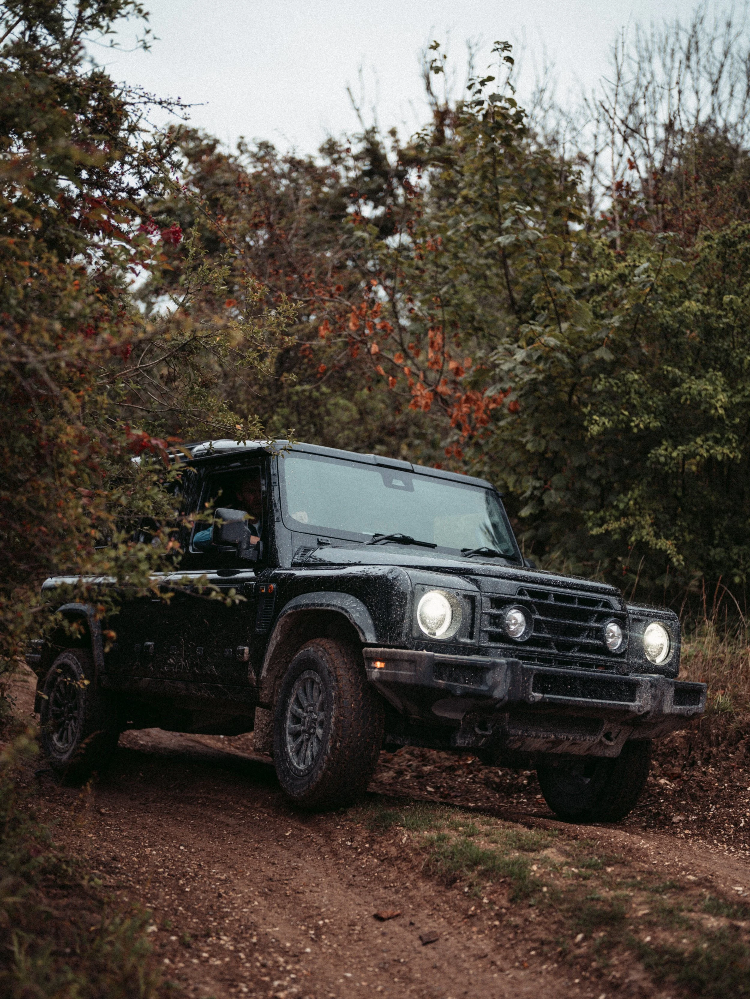 A black off-road vehicle driving on a dirt trail through a forested area with trees and bushes, some with colorful leaves, under a cloudy sky.