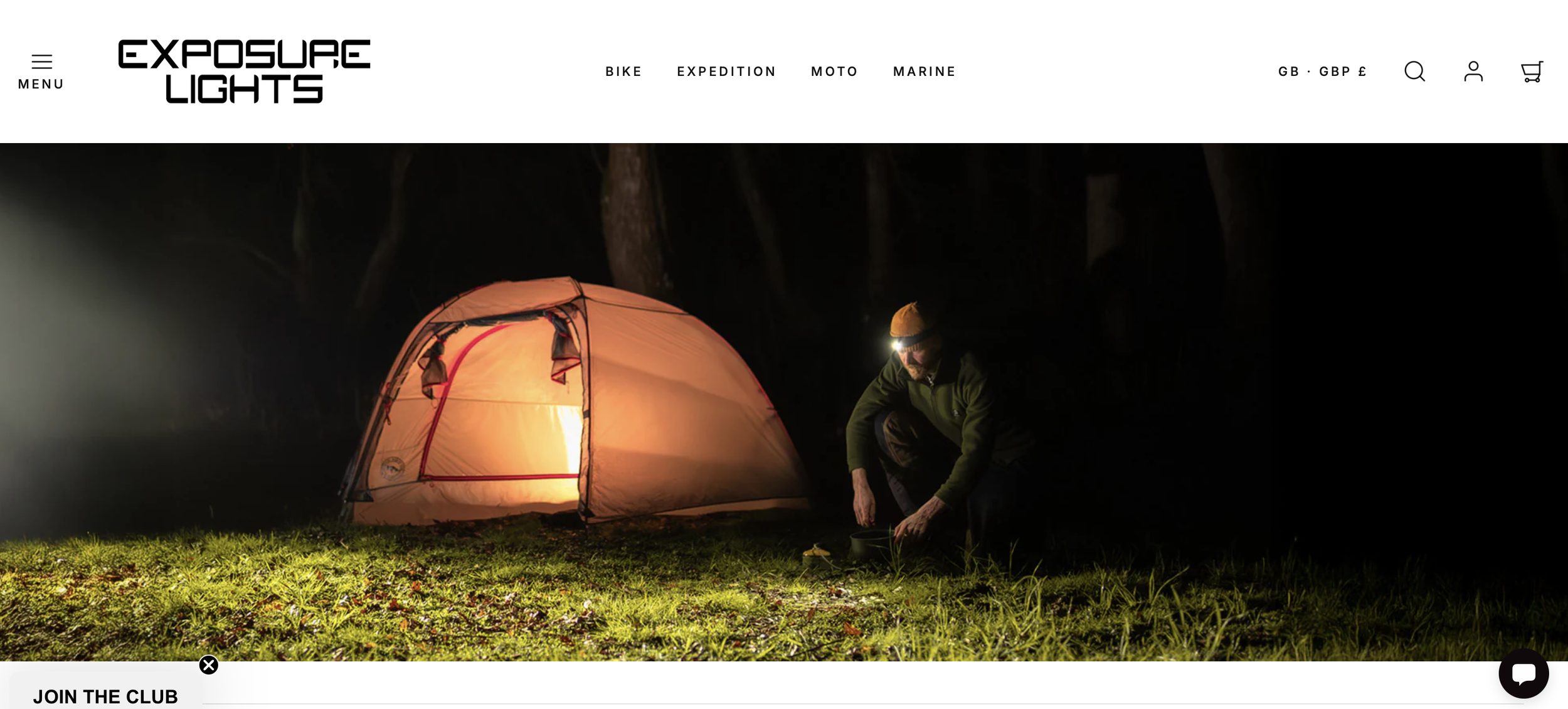 A person wearing a headlamp shaping soil next to a lit camping tent at night in a forest.