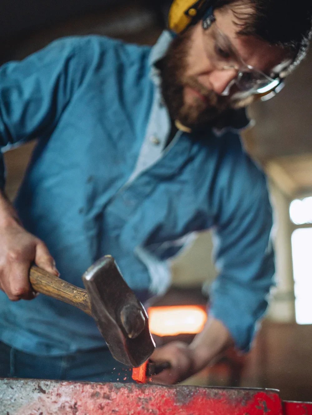 A man with glasses, a beard, and a yellow hard hat hammering on a metal piece, sparks flying, indoors with a window in the background.
