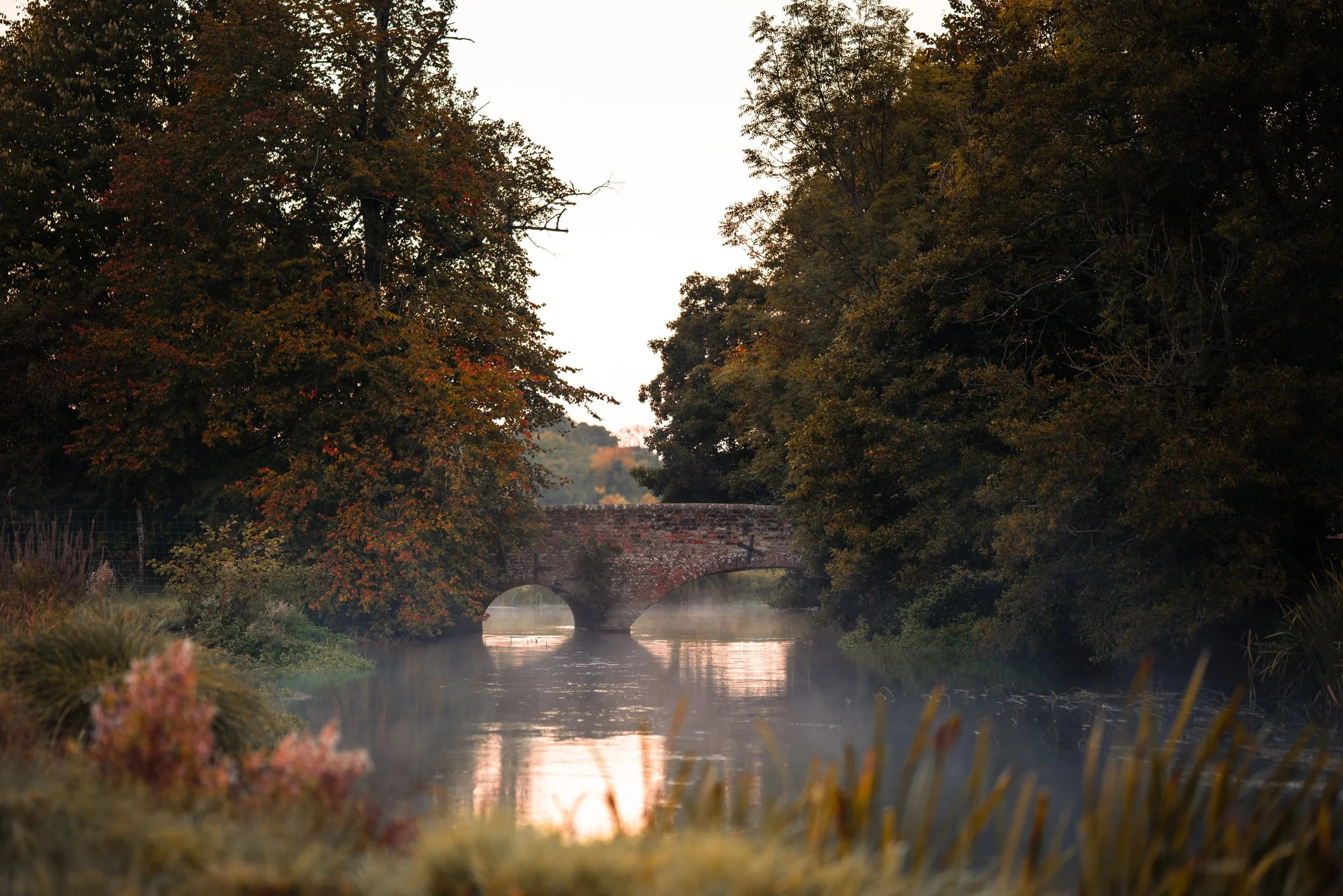 Autumn scene of a brick bridge over a river, surrounded by trees with colorful fall foliage.