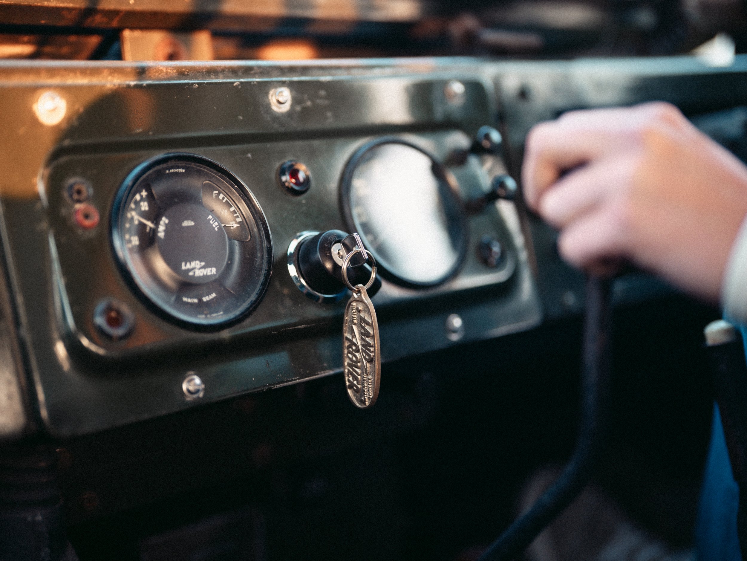 Close-up of a vintage Land Rover dashboard with a person turning the ignition key, which has a Land Rover keychain attached, inside a vehicle.