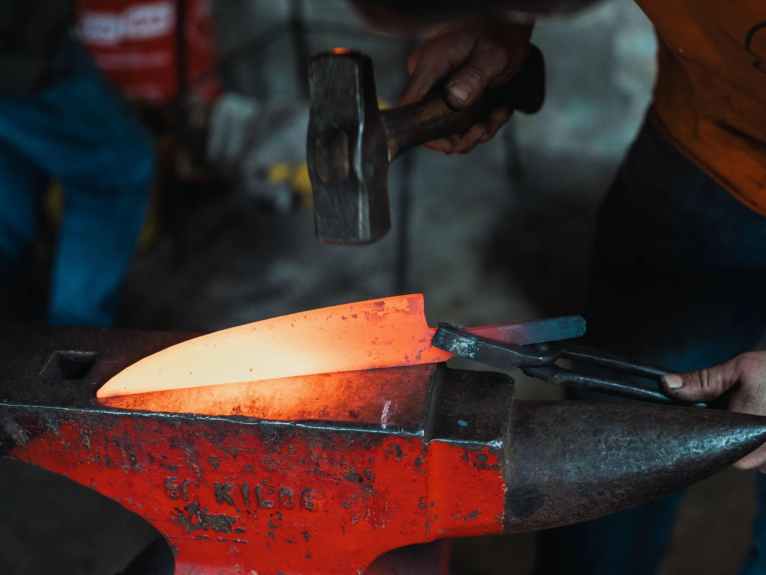 A person heating metal on a red forging forge, with glow from the heated metal illuminating the scene.