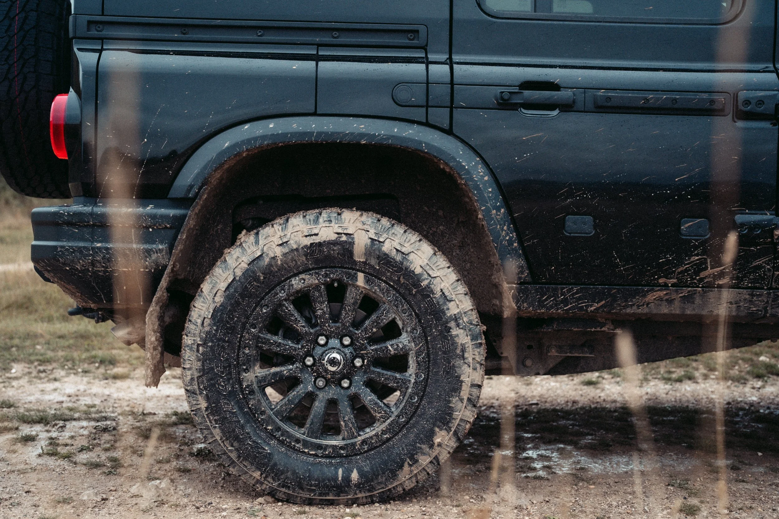 Close-up of a black off-road vehicle with muddy tires, dirt splashed on the wheel well, and a red taillight visible on the left side.