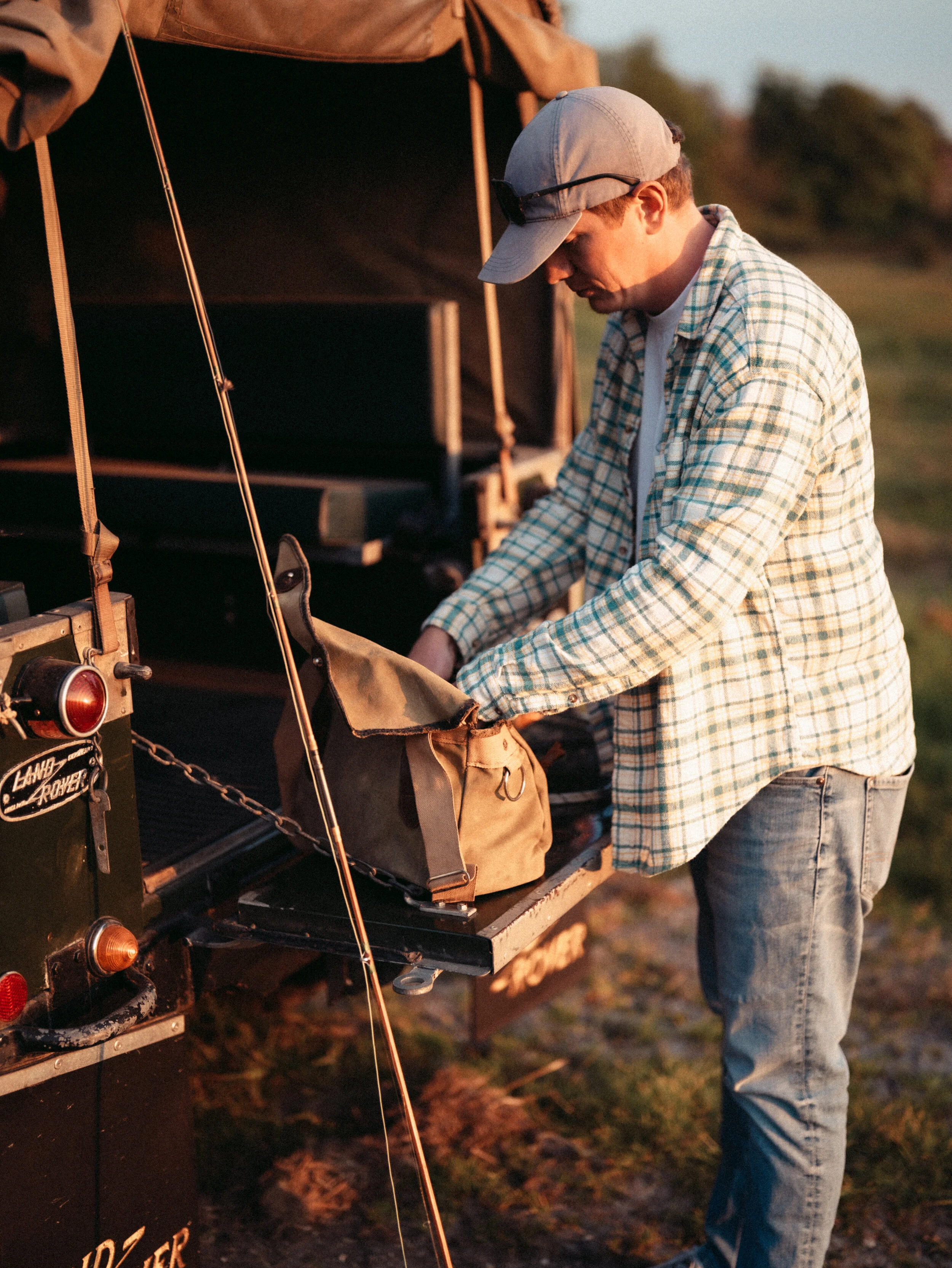A young man wearing a gray cap, plaid shirt, and jeans stands outdoors during sunset, handling equipment on a black camping or expedition vehicle with orange lights and gear attached to it.