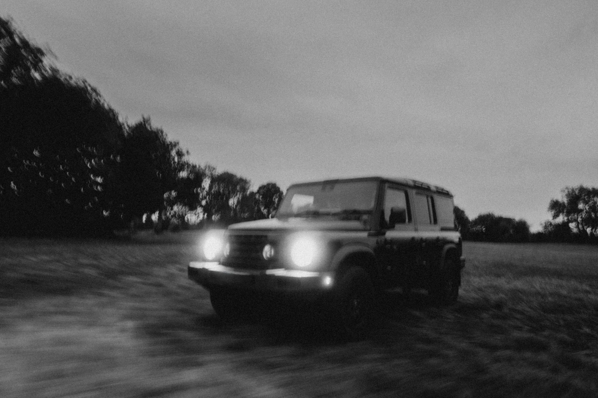 Black and white photo of an SUV driving on a road at night, with trees and a clear sky in the background.