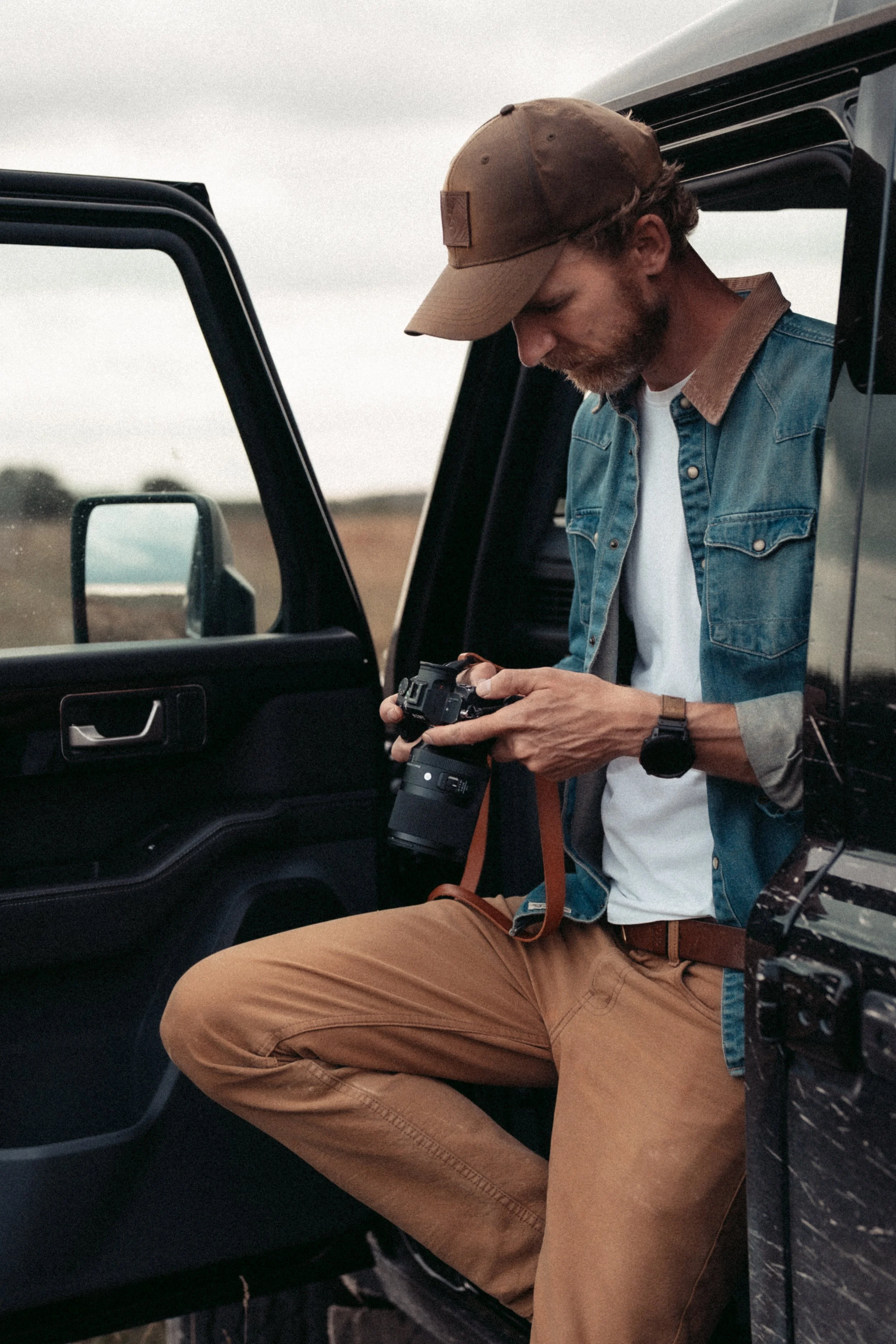 A man with a beard, wearing a beige cap, denim jacket with brown collar, and khaki pants, sitting in the back of a vehicle, looking at a camera in his hands.