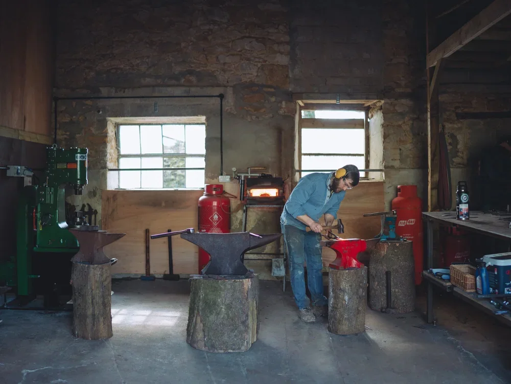 A man in a workshop hammering a piece of metal held on an anvil with a red base, surrounded by other anvils and tools, in a rustic space with brick walls and two windows.