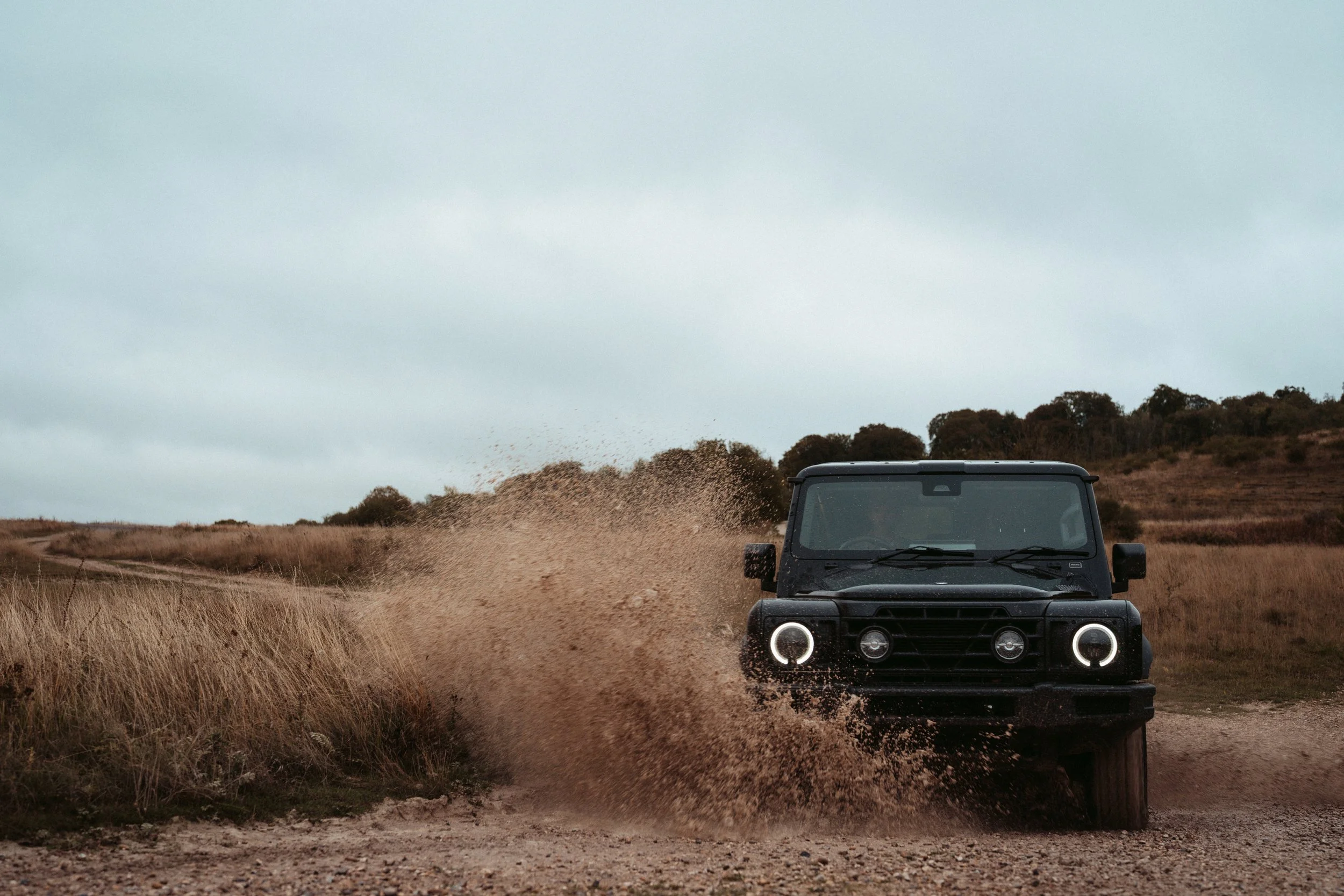 A black off-road vehicle driving through a muddy trail in a rural area with grass and trees under a cloudy sky.