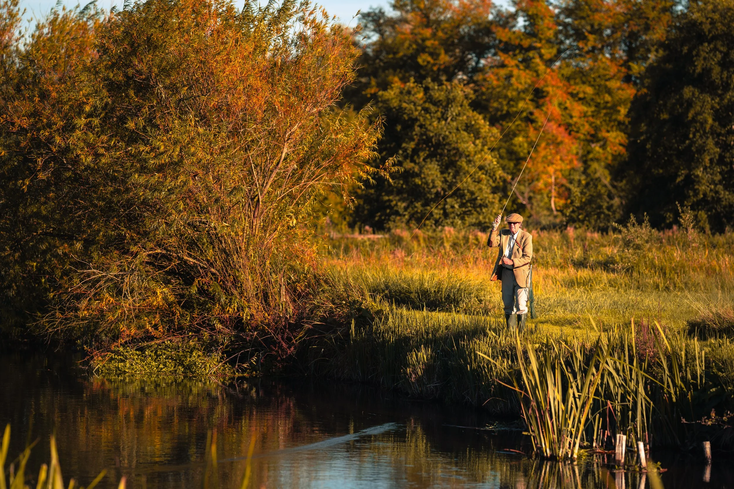 A man walking with a fishing rod beside a river in a park during autumn, with trees displaying fall foliage.