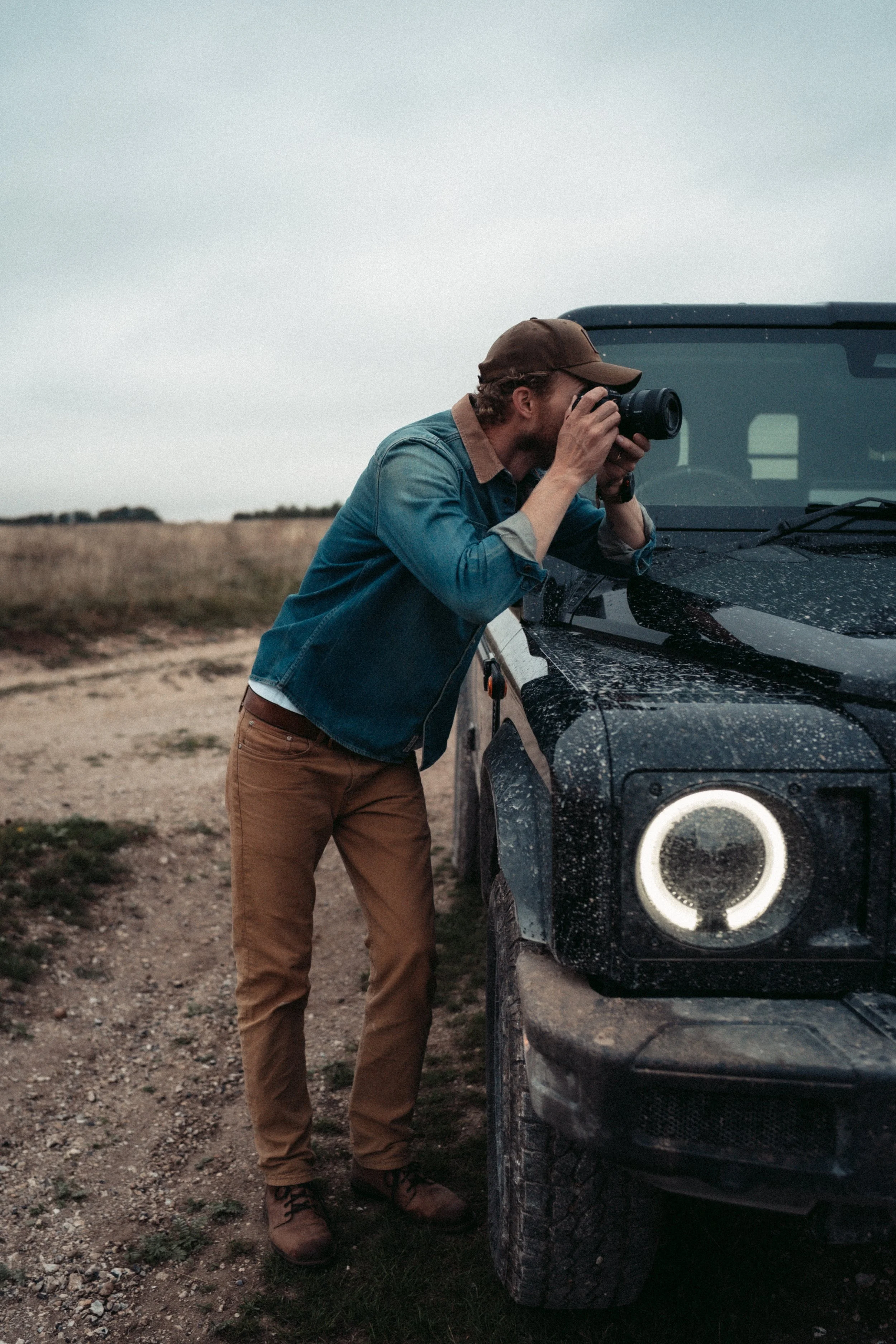A man in a denim jacket, brown pants, and a baseball cap is leaning over the front of a black off-road vehicle, looking through a camera. The vehicle is muddy, and the man appears to be taking a photo or inspecting something near the car's hood. The 