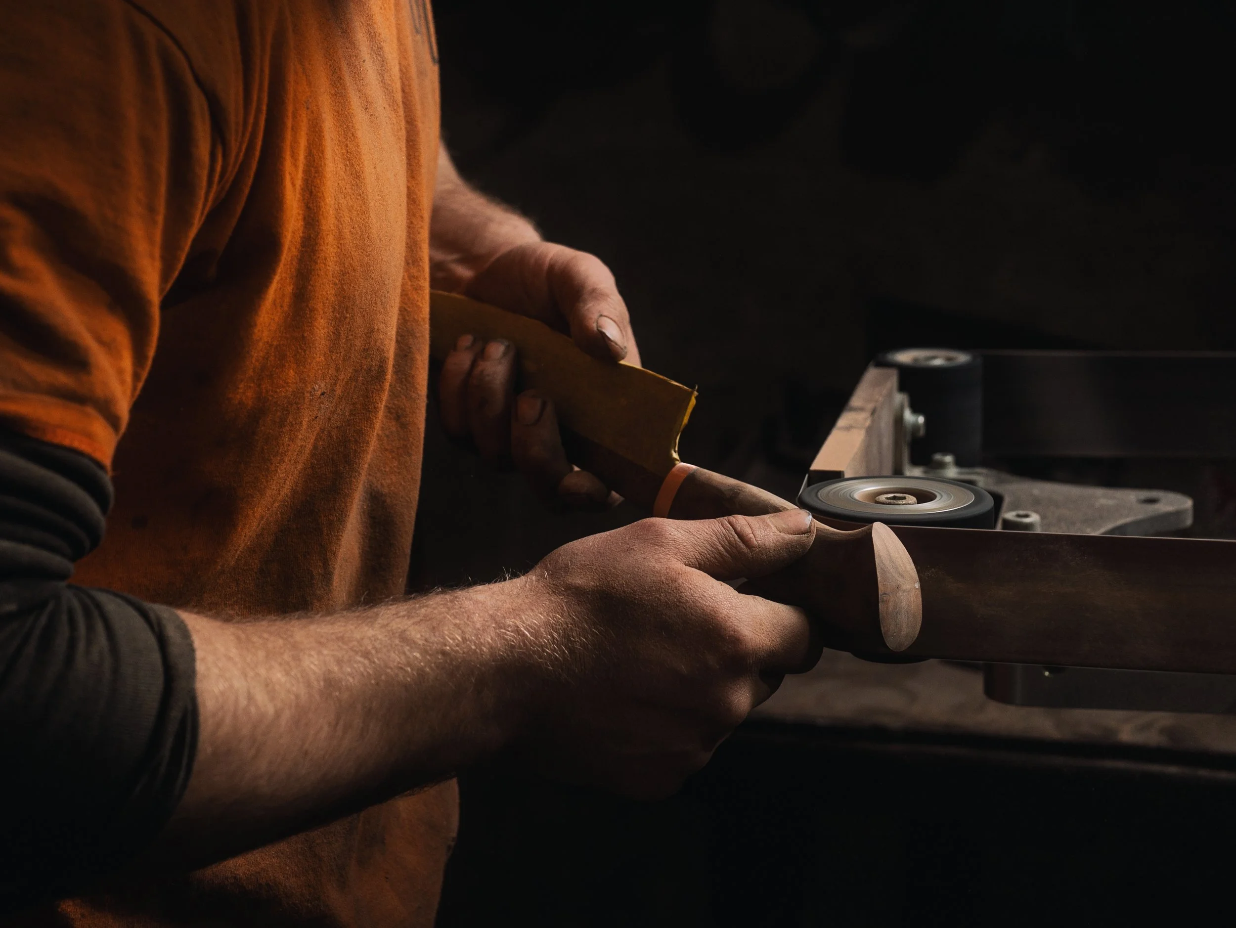 Close-up of a person's hands holding a tool while working on a piece of wood, with a grinding or polishing machine in the background.