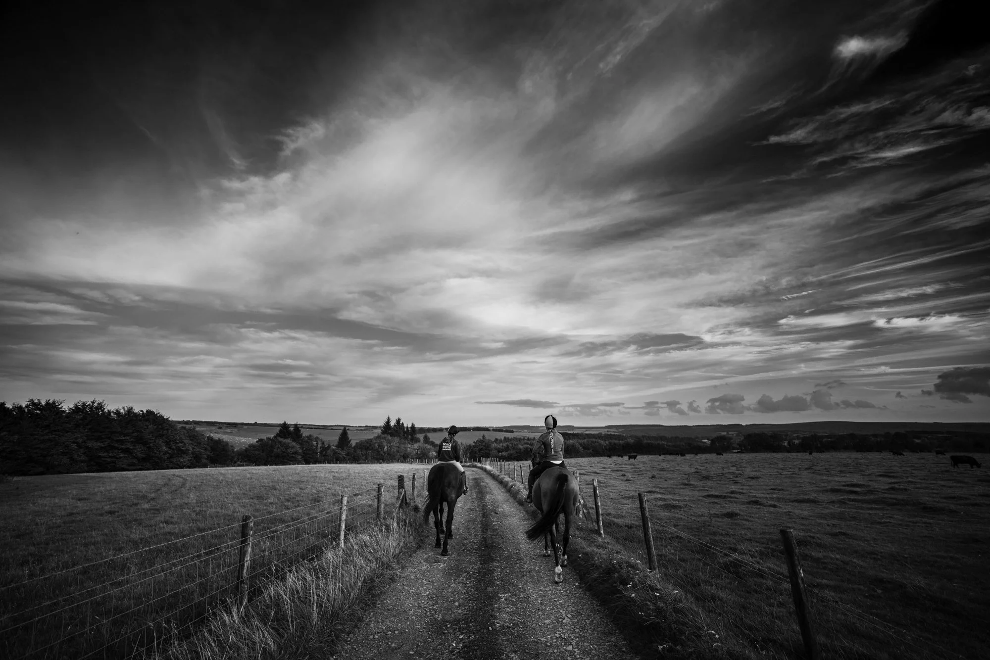 Two people riding horses down a rural dirt path bordered by a fence, with open fields and a cloudy sky in black and white.