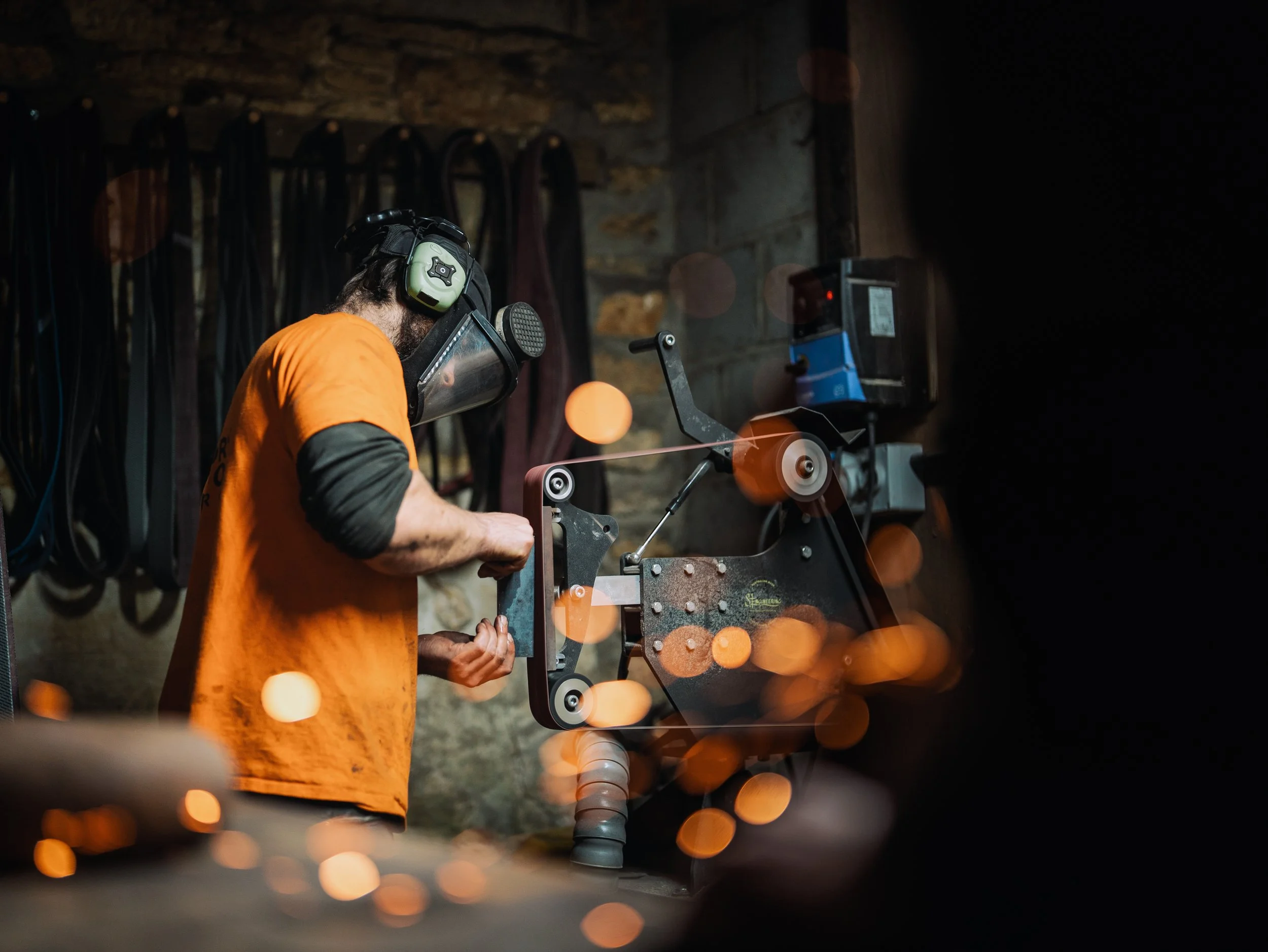 A man operating a metalworking or crafting machine, wearing safety ear protection and an orange shirt, in a dimly lit workshop with various straps or belts hanging on the wall.
