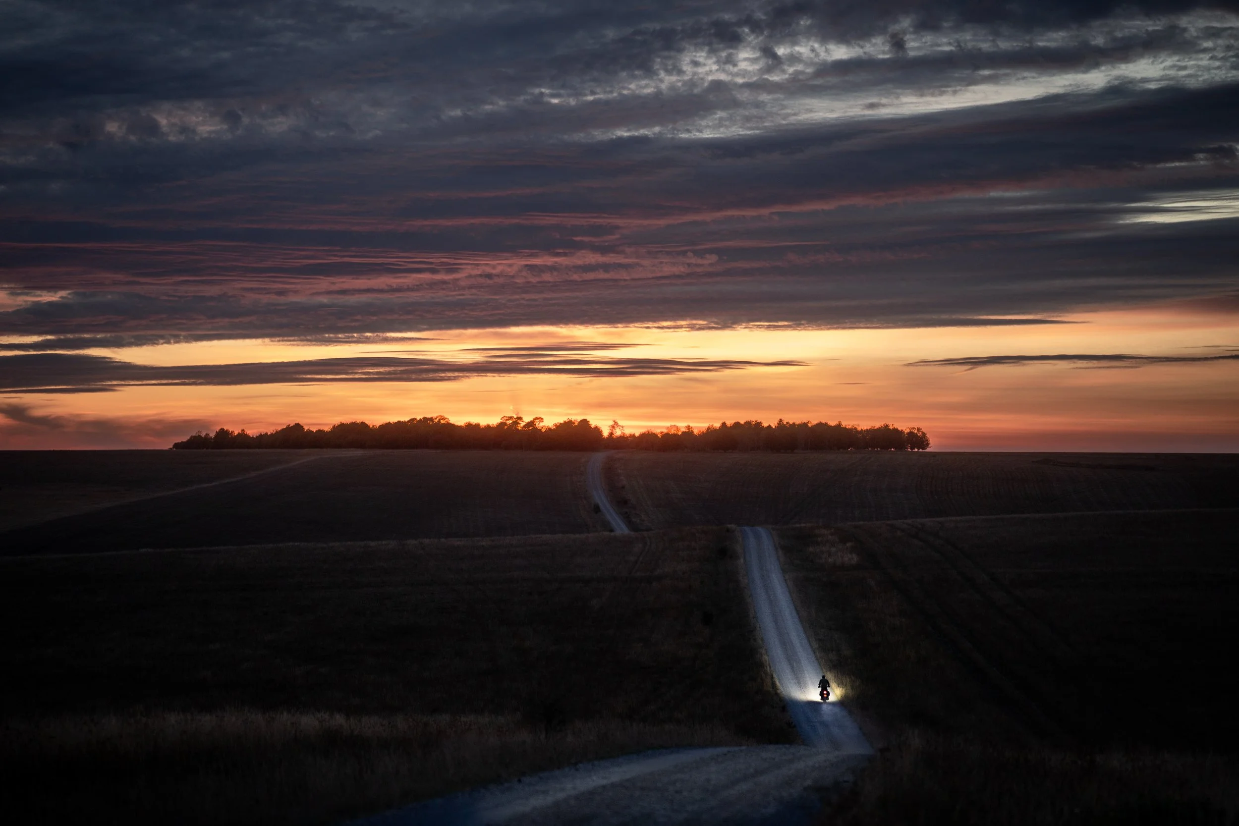 A sunset with colorful clouds across the sky and a faraway tree line on the horizon. A motorcycle with a bright light is riding down a winding road through an open field.