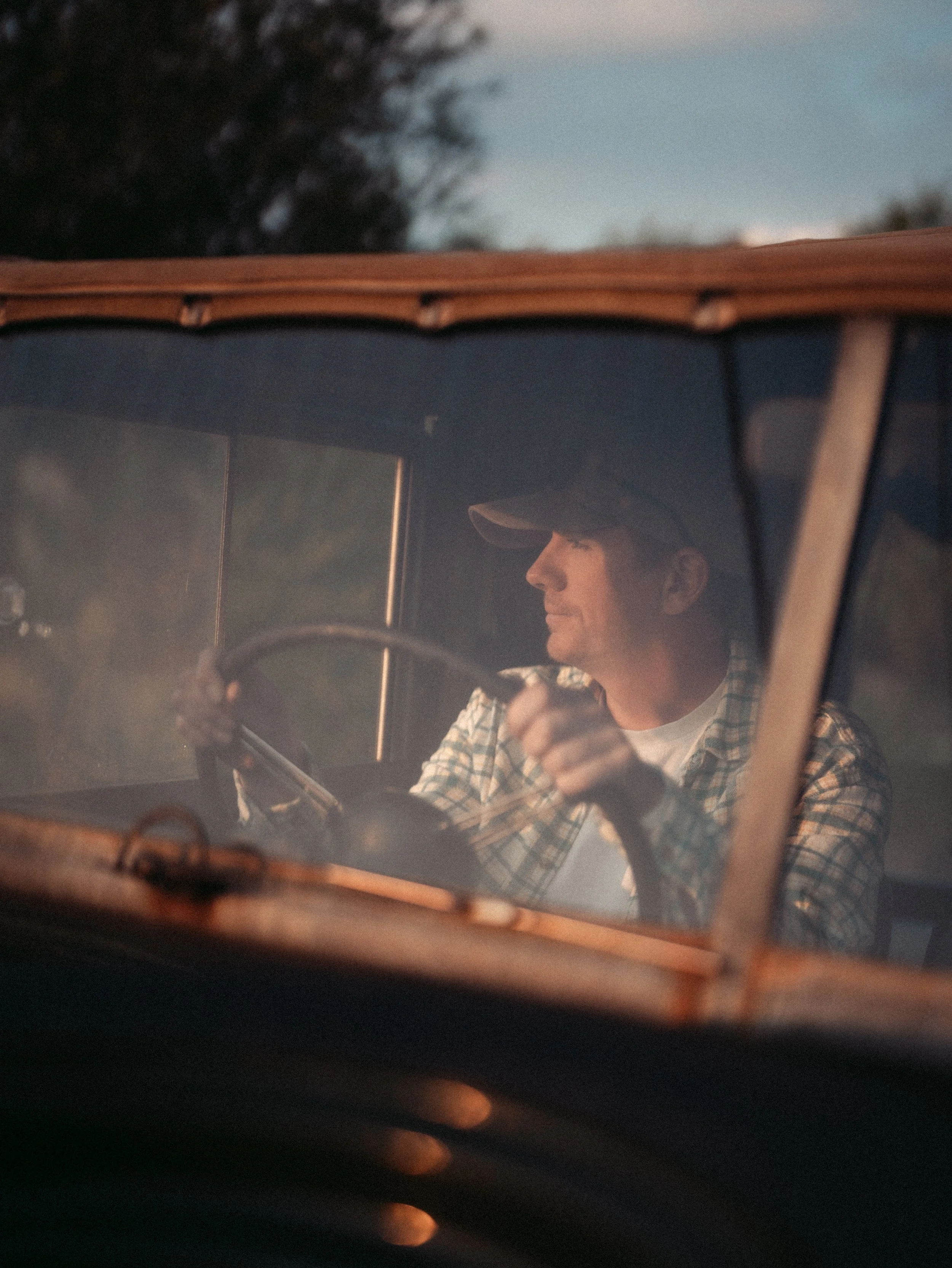A man wearing a plaid shirt and baseball cap is driving a vintage truck, viewed from outside through the windshield during sunset or sunrise.
