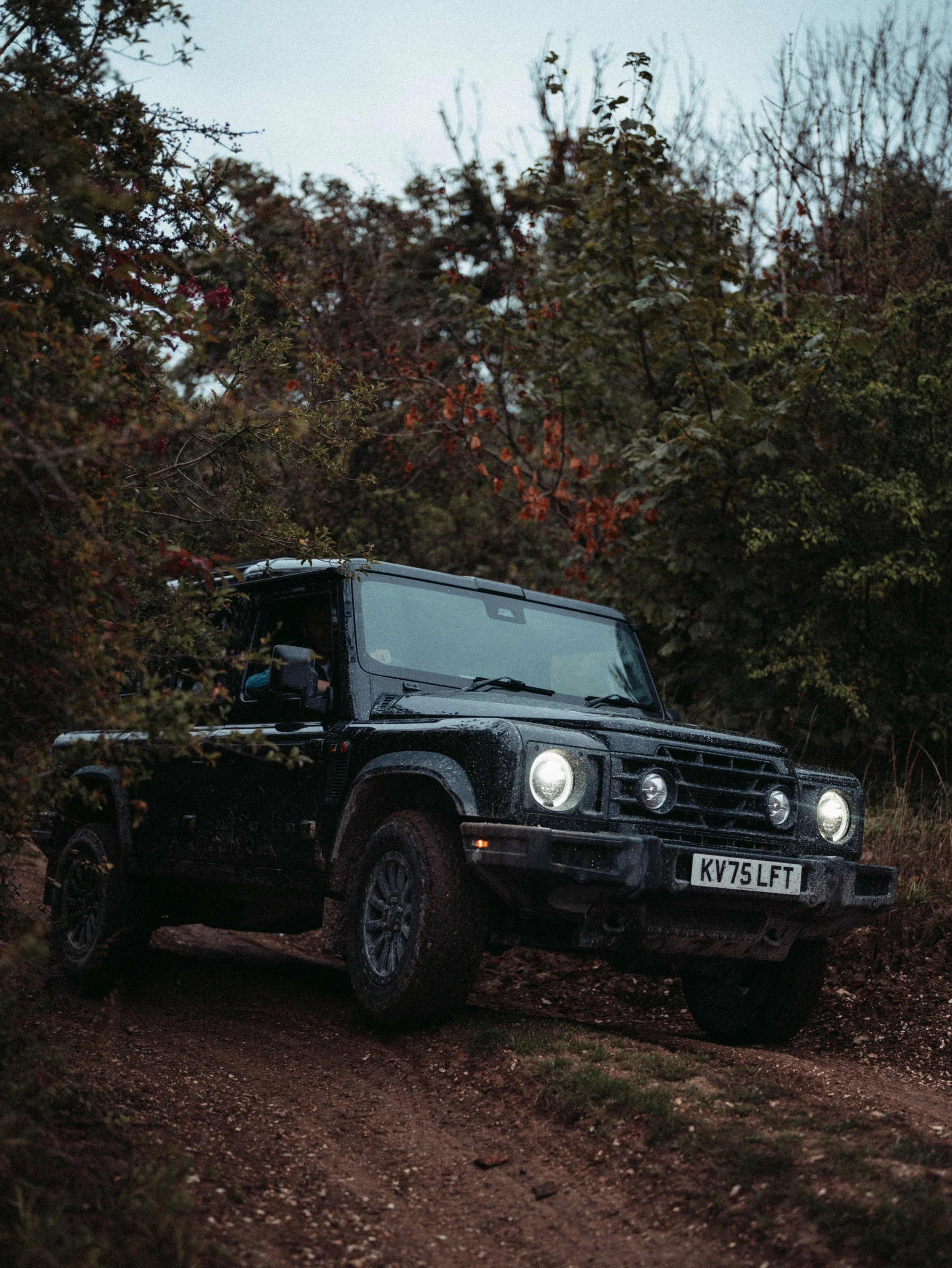 A black off-road vehicle with headlights on, parked on a dirt path surrounded by dense foliage, including trees and bushes, during dusk or early evening.