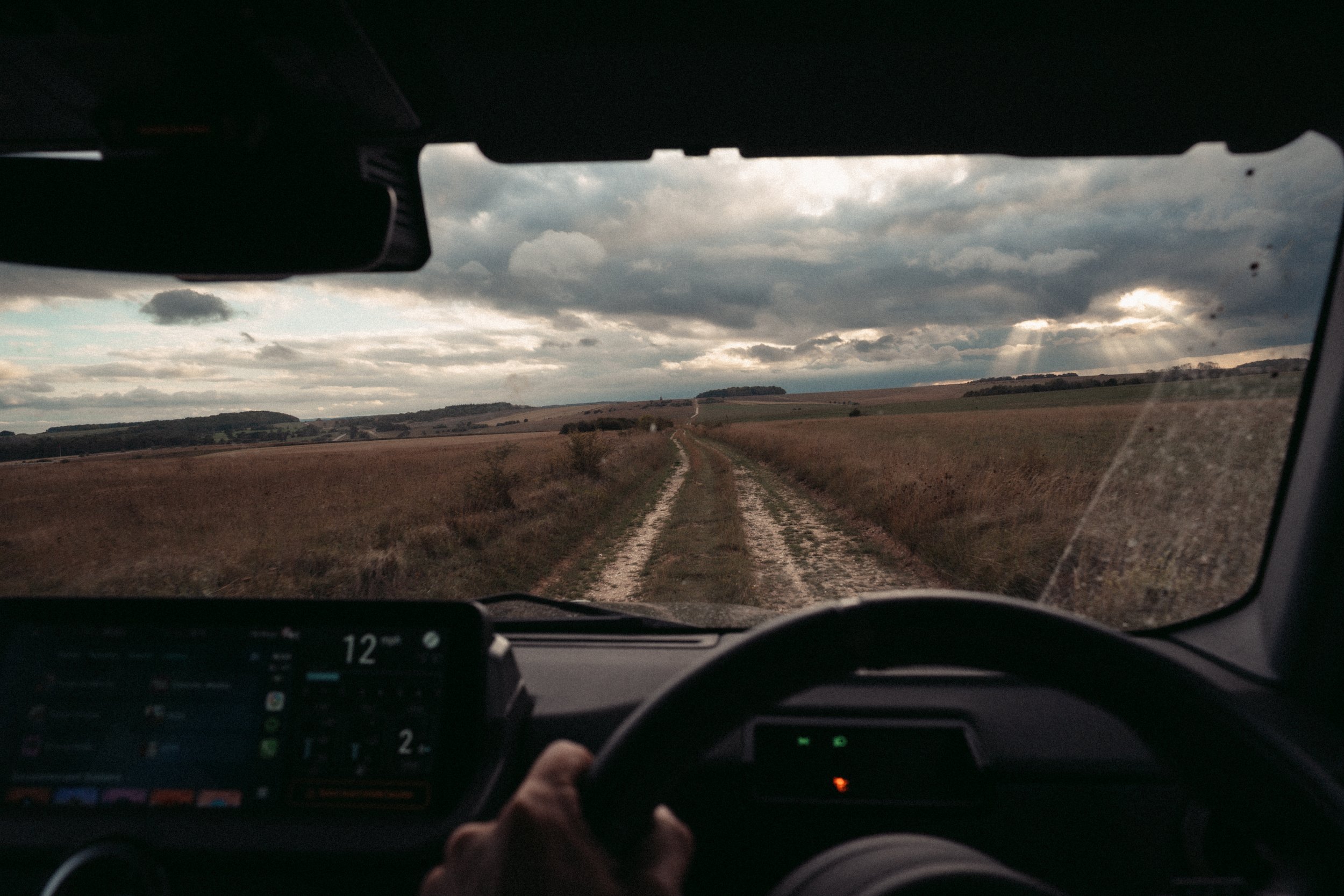 Inside a vehicle, looking through the windshield at a dirt road stretching into the distance across open fields with cloudy sky overhead.