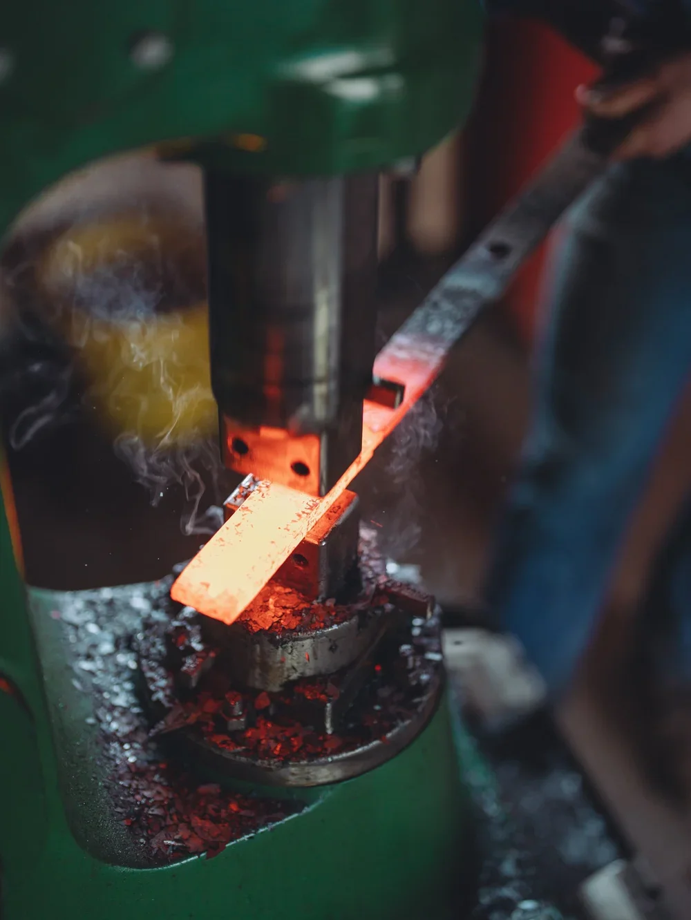 A metal piece being forged in a blacksmithing press with sparks and glowing red heat.
