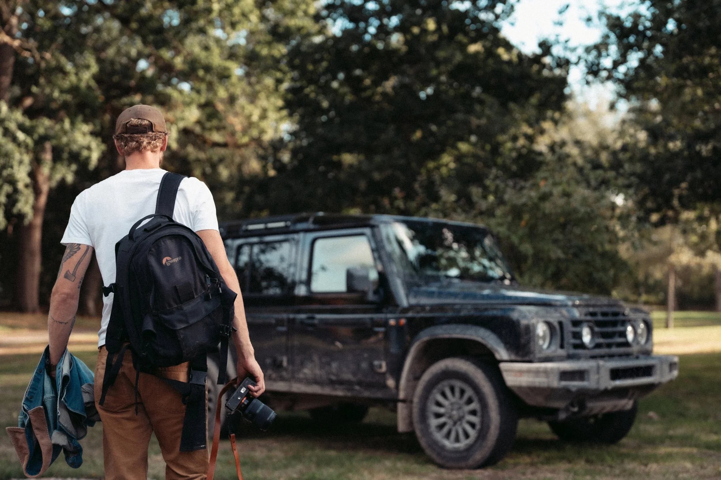 Back view of a young man with curly blond hair wearing a brown cap, white T-shirt, and brown pants, carrying a black backpack and a rolled-up blue jacket, holding a camera, walking past a black SUV in a park with green trees.