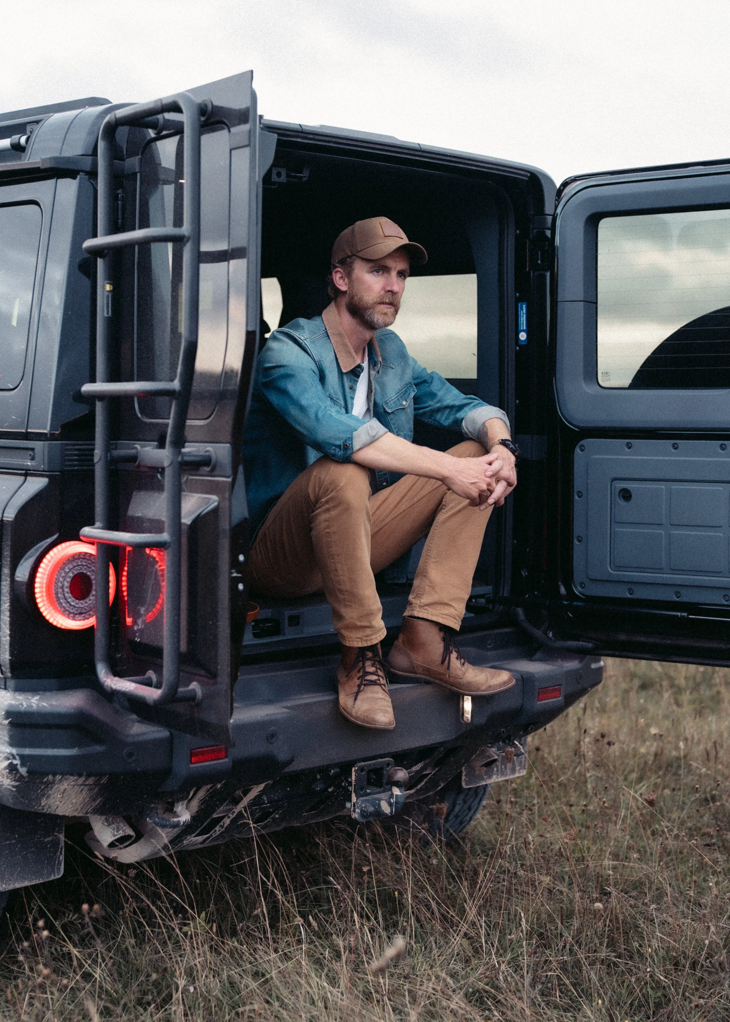 A man with a beard and brown hair sitting on the back of a black off-road vehicle in a grassy field during daytime, wearing a brown cap, denim shirt, and brown pants, looking contemplative.
