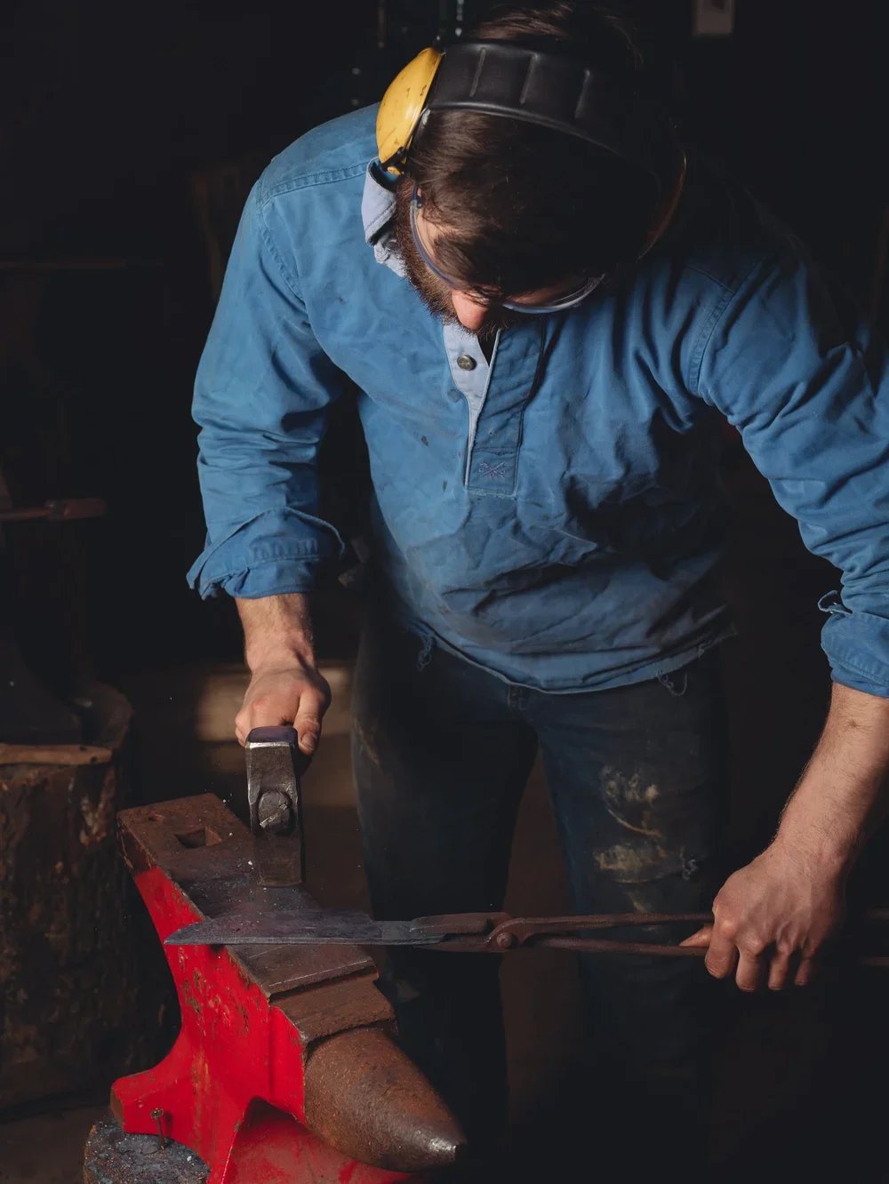 Man using a hammer and chisel on a heated piece of metal on an anvil, wearing ear protection and a blue shirt.