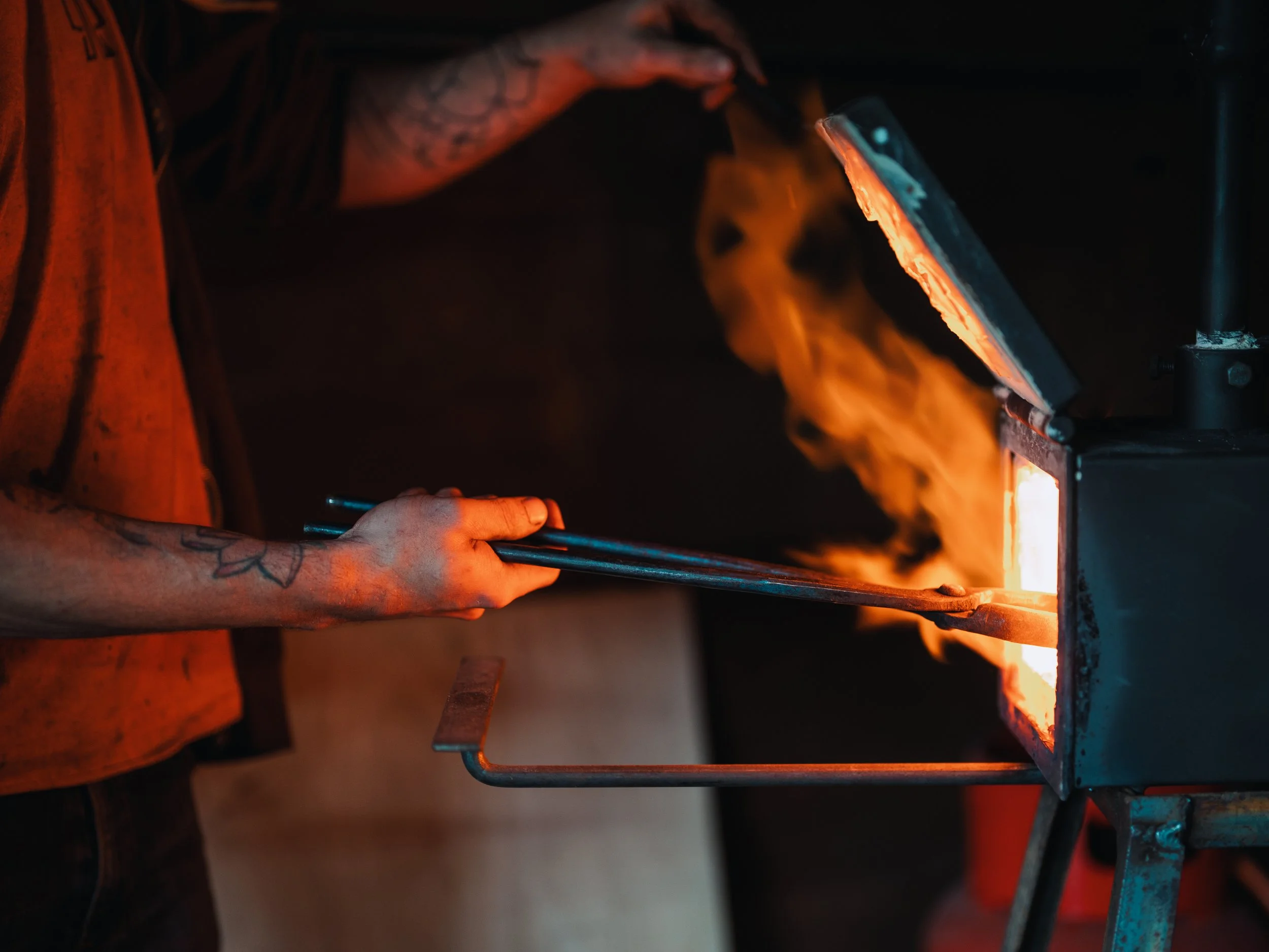 Close-up of tattooed hands adjusting a piece of hot metal in a forge or furnace, with bright orange flames illuminating the scene.