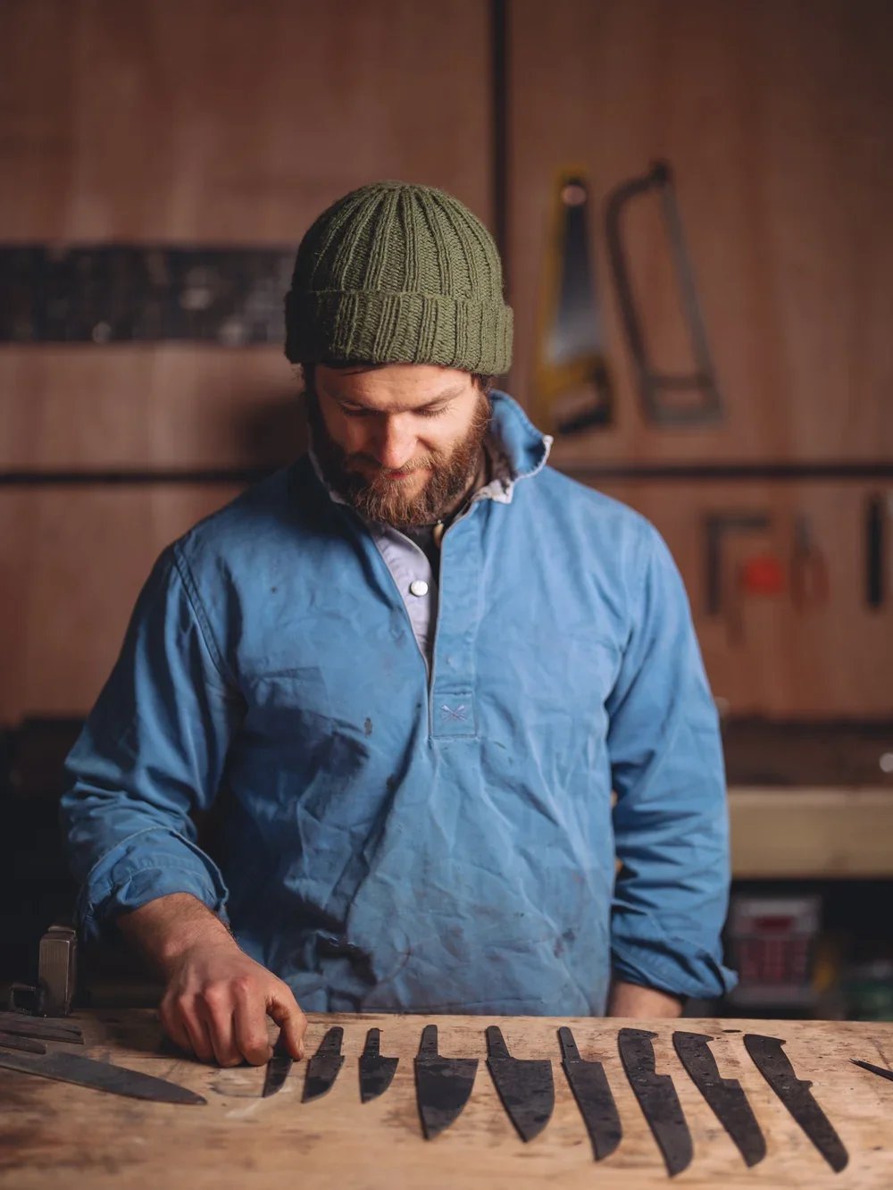 A man with a beard wearing a green beanie and a blue work jacket looking down at a workbench with several wood carving knives.