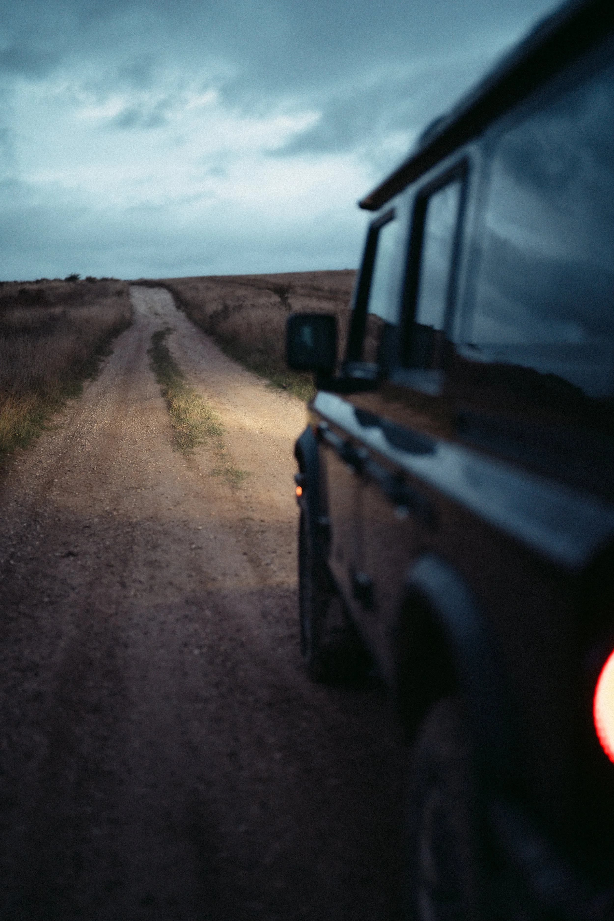 A dirt country road stretching into the horizon under a cloudy sky, with a vehicle partially visible on the right side of the image.