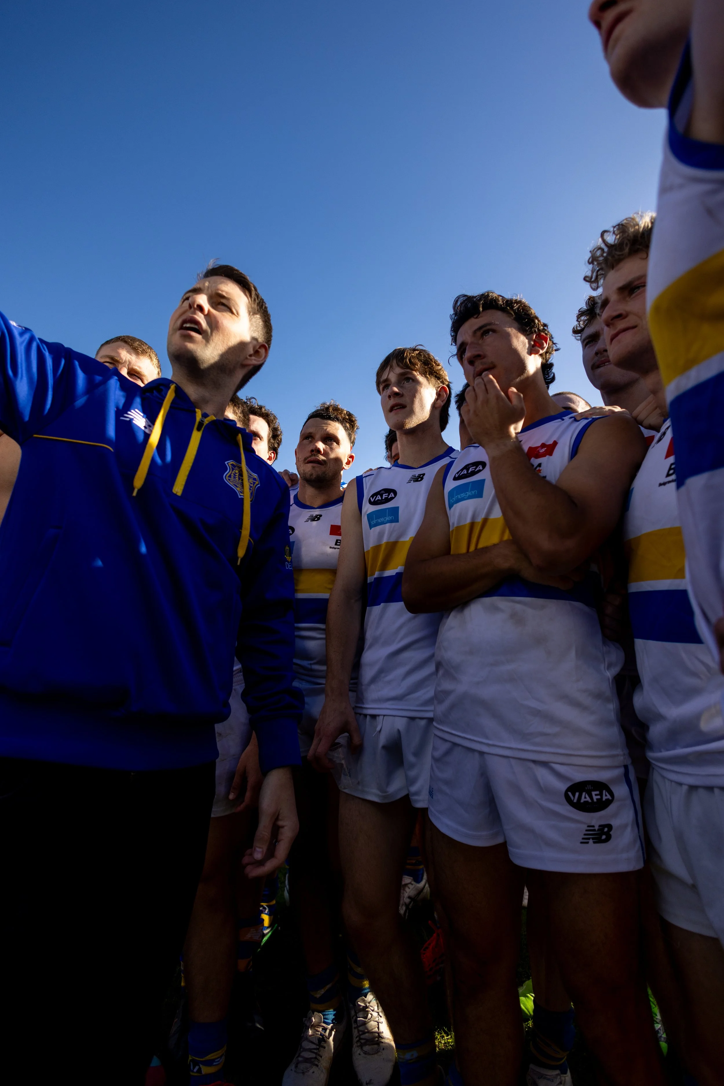 A football team is gathered together during a huddle, wearing white and blue uniforms, with a coach or staff member in a blue jacket speaking to them, against a bright blue sky.