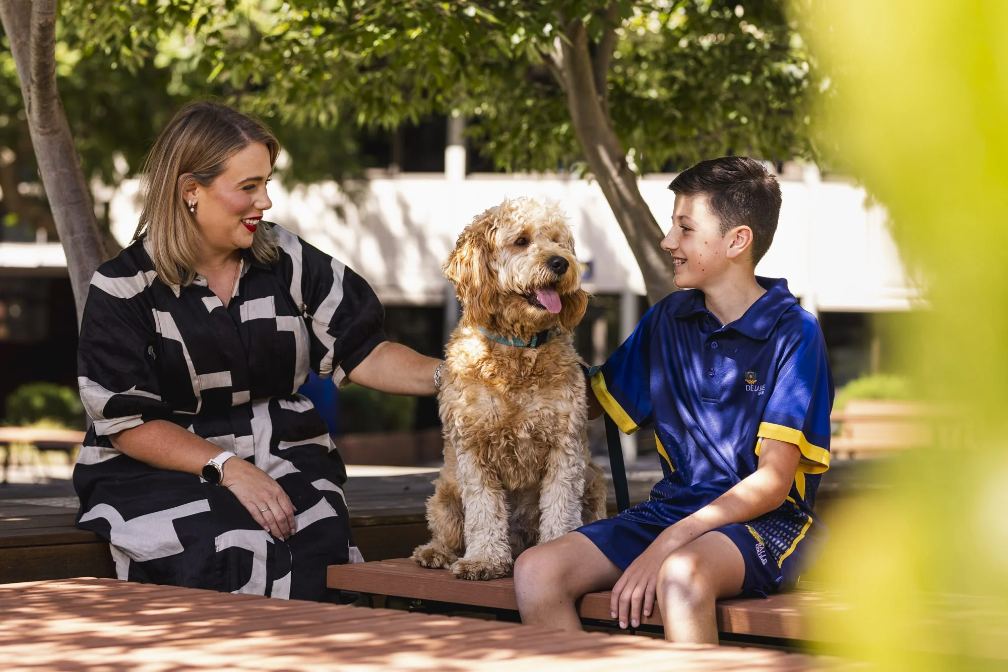 A woman and a boy sitting on a park bench outdoors with a dog between them, smiling and looking at each other.