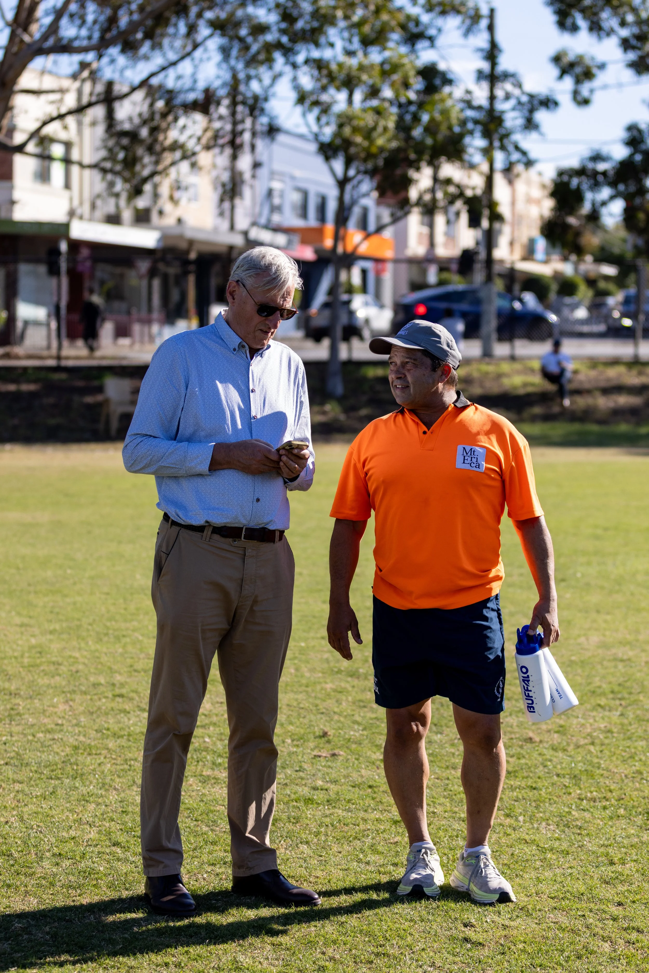Two men are standing on a grassy field having a conversation. One man is dressed casually in beige pants and a light blue button-up shirt, looking at his phone. The other man is wearing a bright orange sports shirt, dark shorts, and a gray cap, holding a water bottle. The background shows trees, buildings, and a few people on a sunny day.