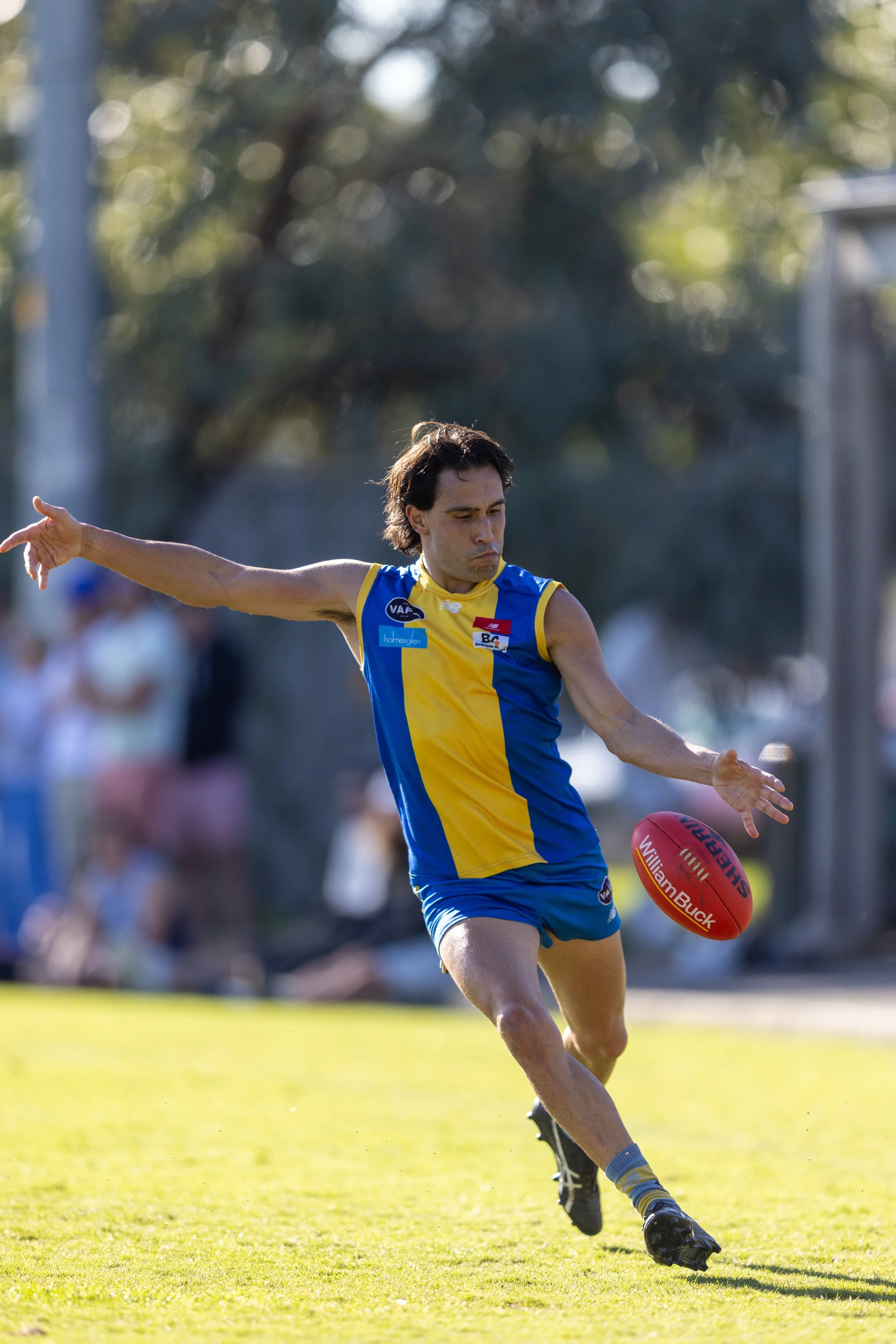 An Australian Rules football player in a blue and yellow uniform kicking a red football on a grassy field.