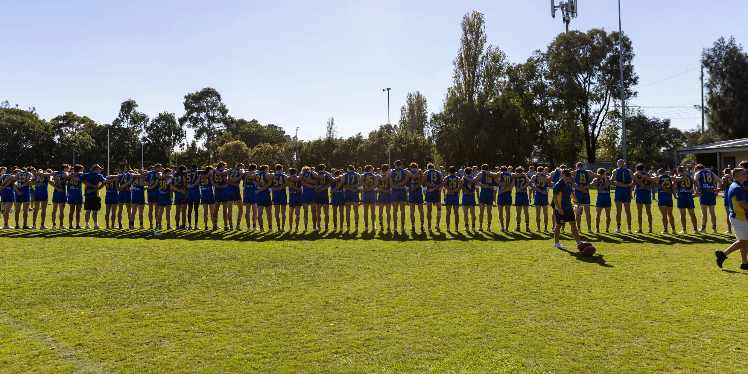 A large group of Australian football players standing in a line with their arms around each other's shoulders on a grassy field, with trees and blue sky in the background.