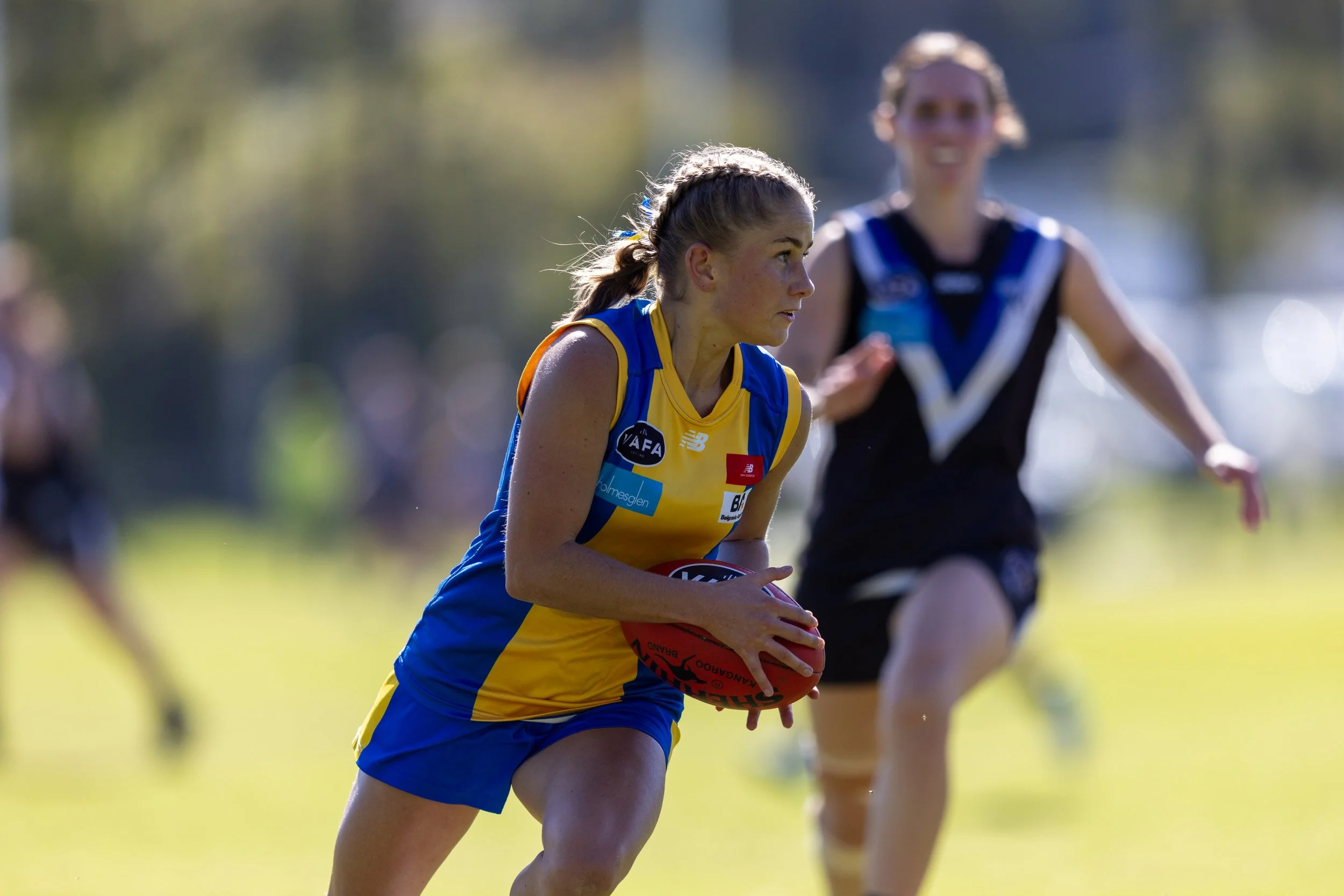 A female athlete in a yellow and blue sports uniform running with a football during a game, with another runner in a black and blue uniform blurred in the background on a grassy field.