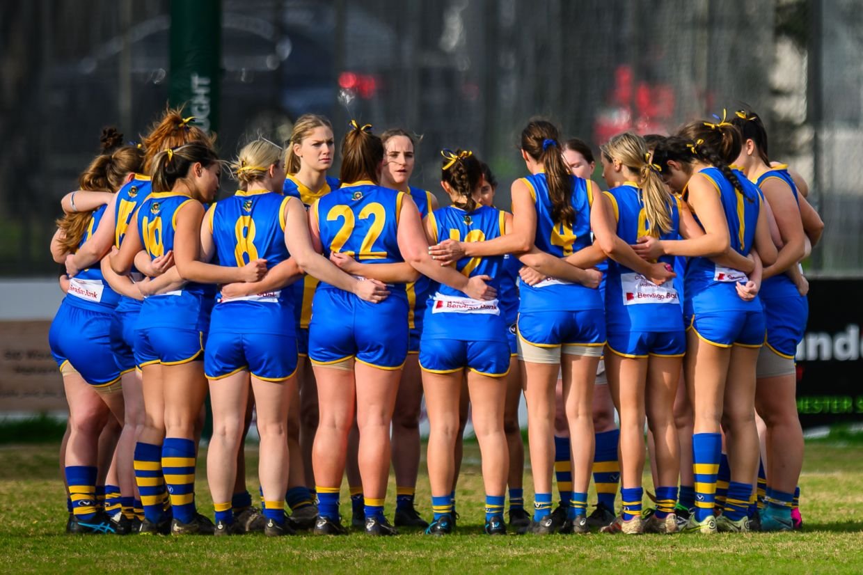 A group of female De La Salle Footballers in blue and yellow sports uniforms huddled together on a field, likely during a team discussion or pre-game pep talk.