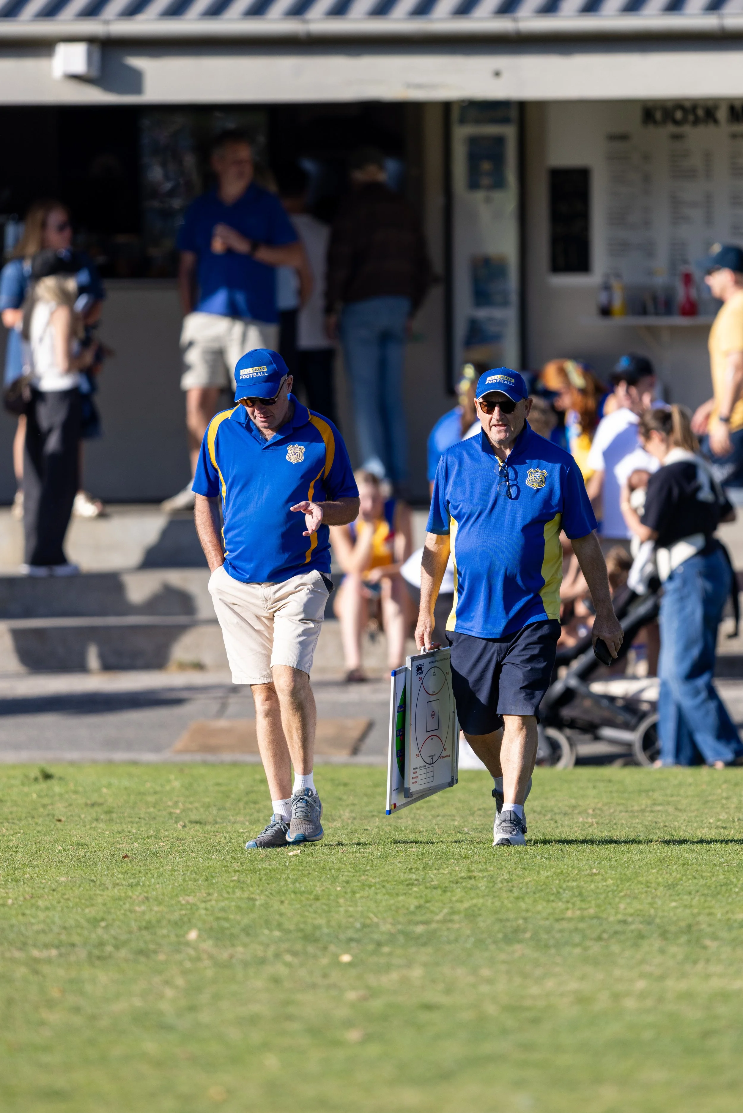 Two De La Salle club men in blue shirts and caps with a sports logo are walking on a grassy field, carrying a hockey tactics board, with people and a food stand in the background.