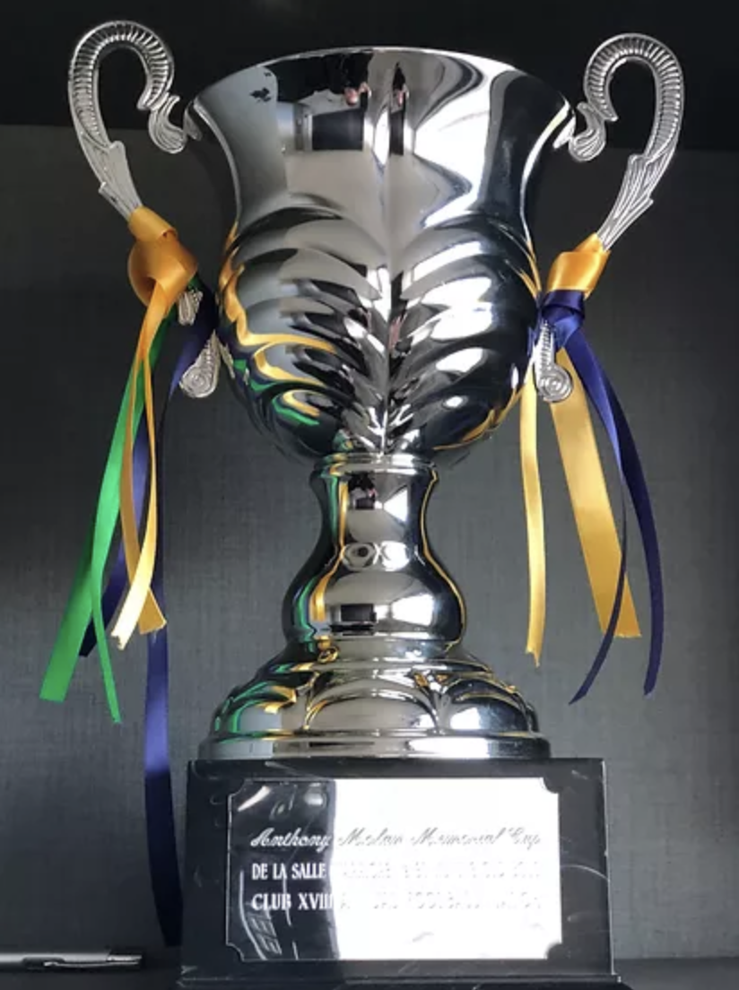 A shiny silver trophy cup with two handles, decorated with yellow, blue, and green ribbons, placed on a black base with a plaque.
