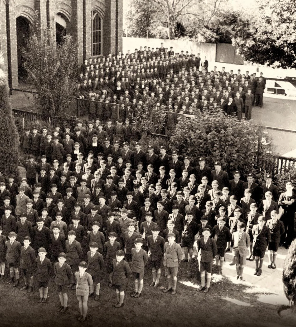 Black-and-white photo of a large school group of boys in suits and hats from De La Salle College, standing outdoors in front of a brick school building with large windows and trees, arranged in rows.