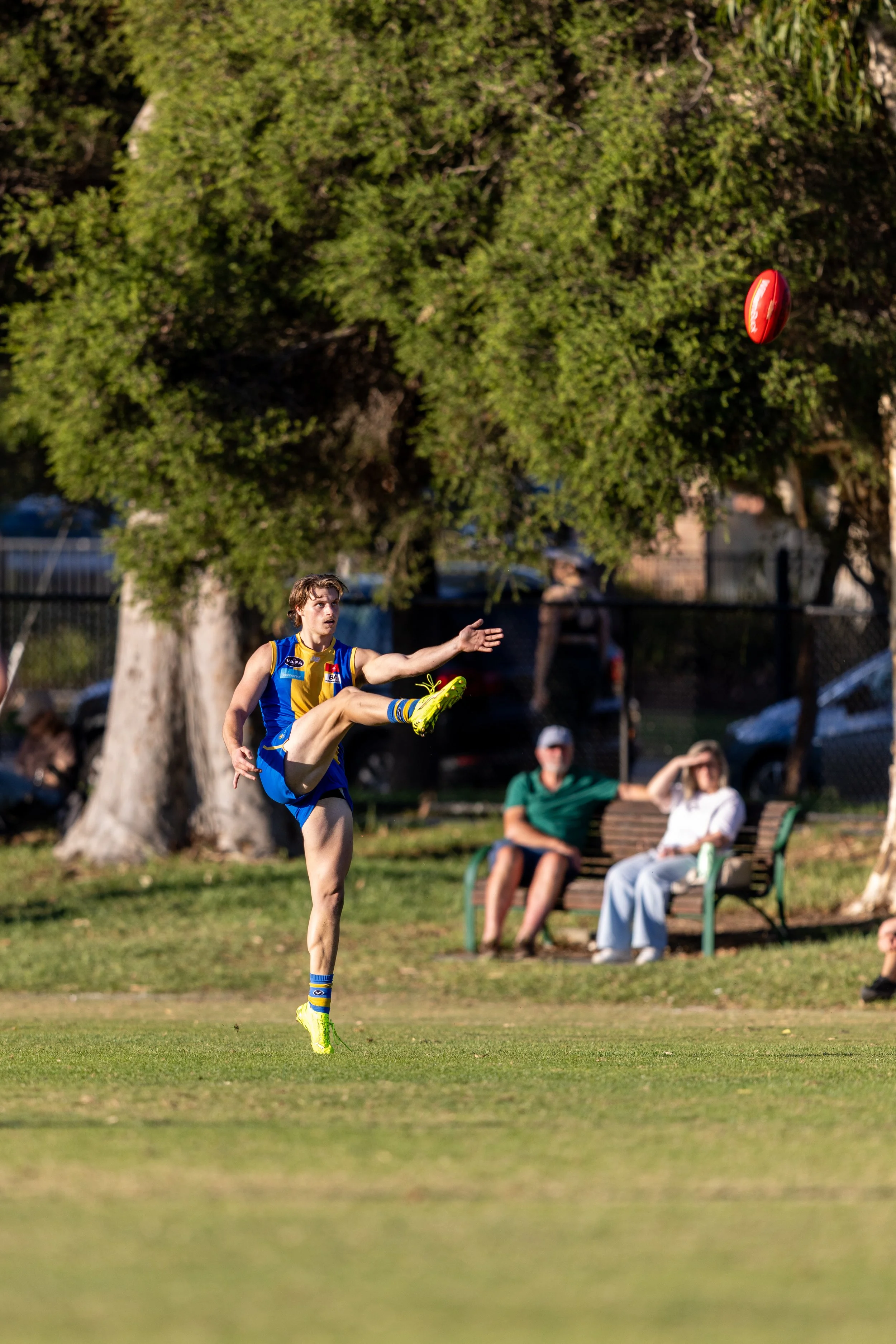 A young man playing Australian rules football outdoors in a park, mid-air, about to kick a red football. He is wearing a blue and yellow sports uniform, yellow shoes, and socks, with a few spectators sitting on a bench in the background.