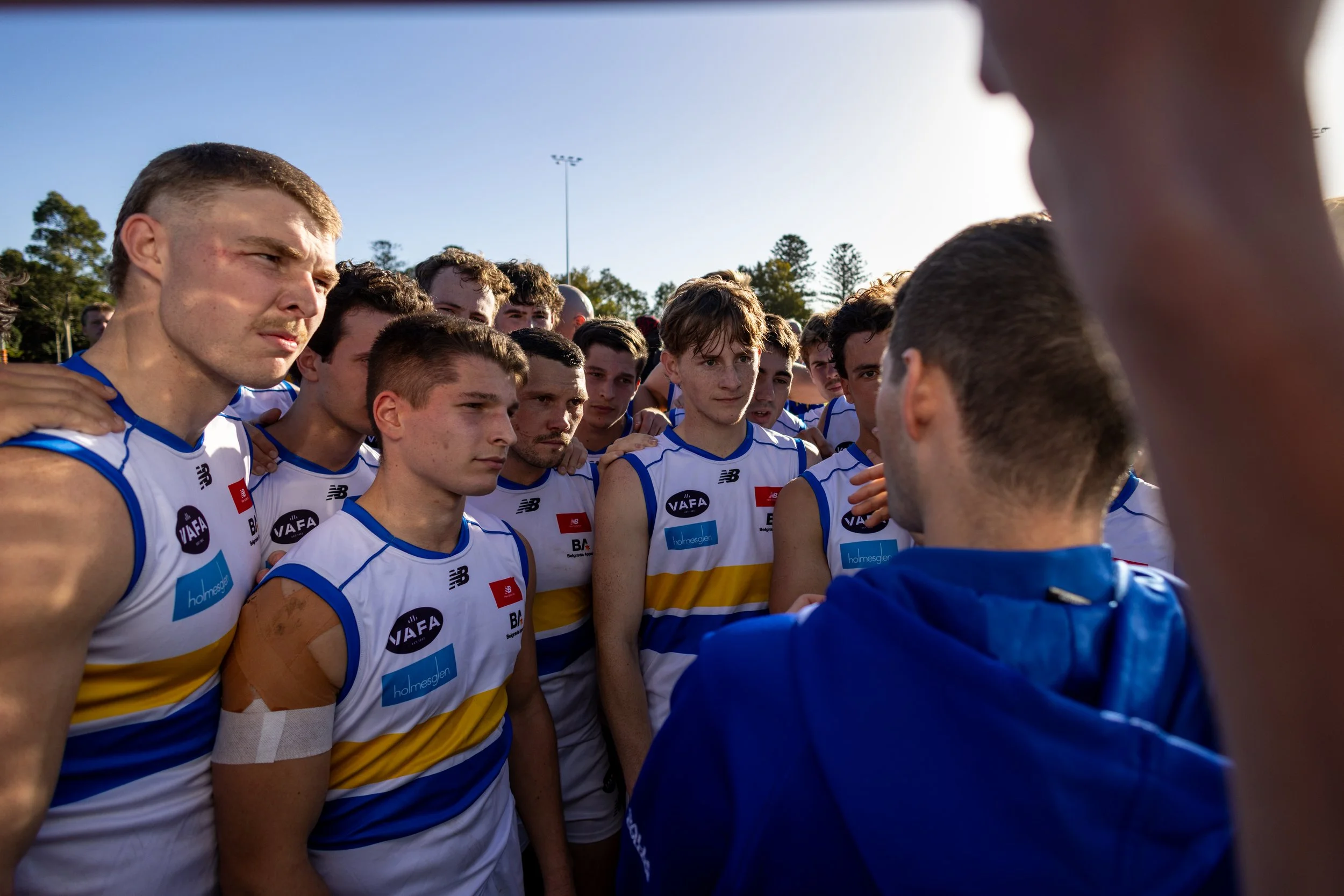 A group of male De La Salle Footballers wearing white and yellow sports uniforms with blue accents, standing close together in a huddle on an outdoor sports field, listening attentively to a coach or team leader whose back is to the camera.