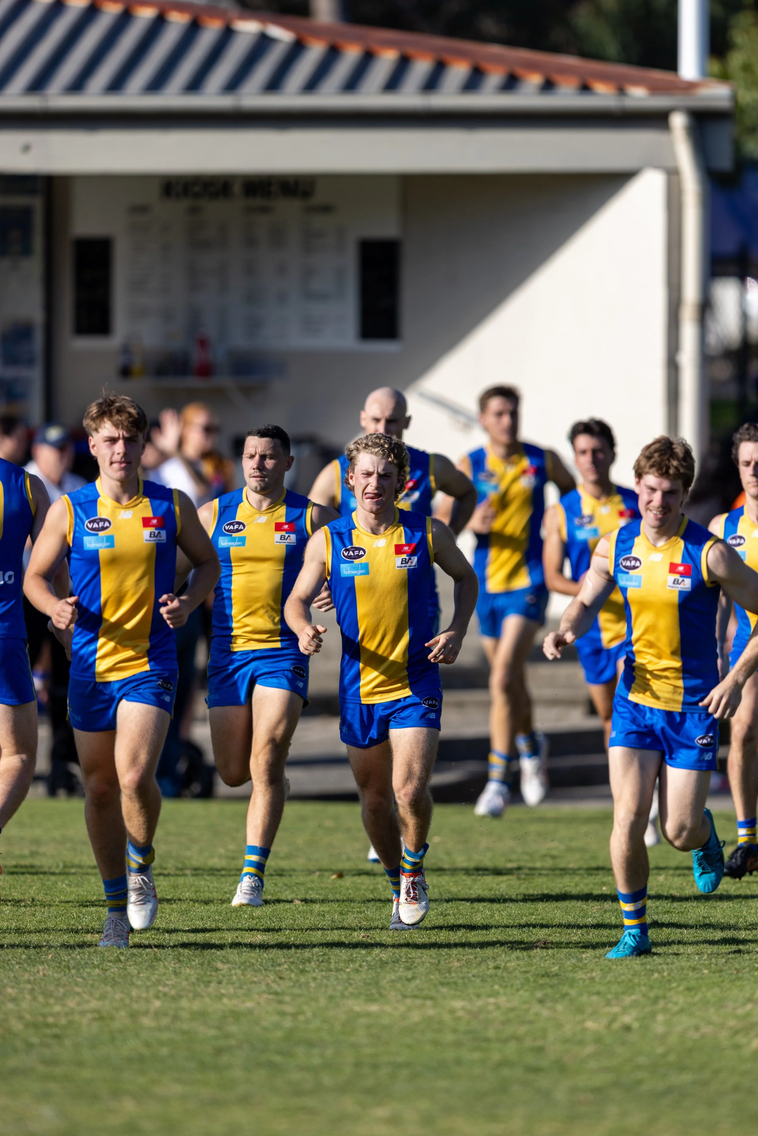 Australian Rules Football team training on the field, wearing blue and yellow uniforms.