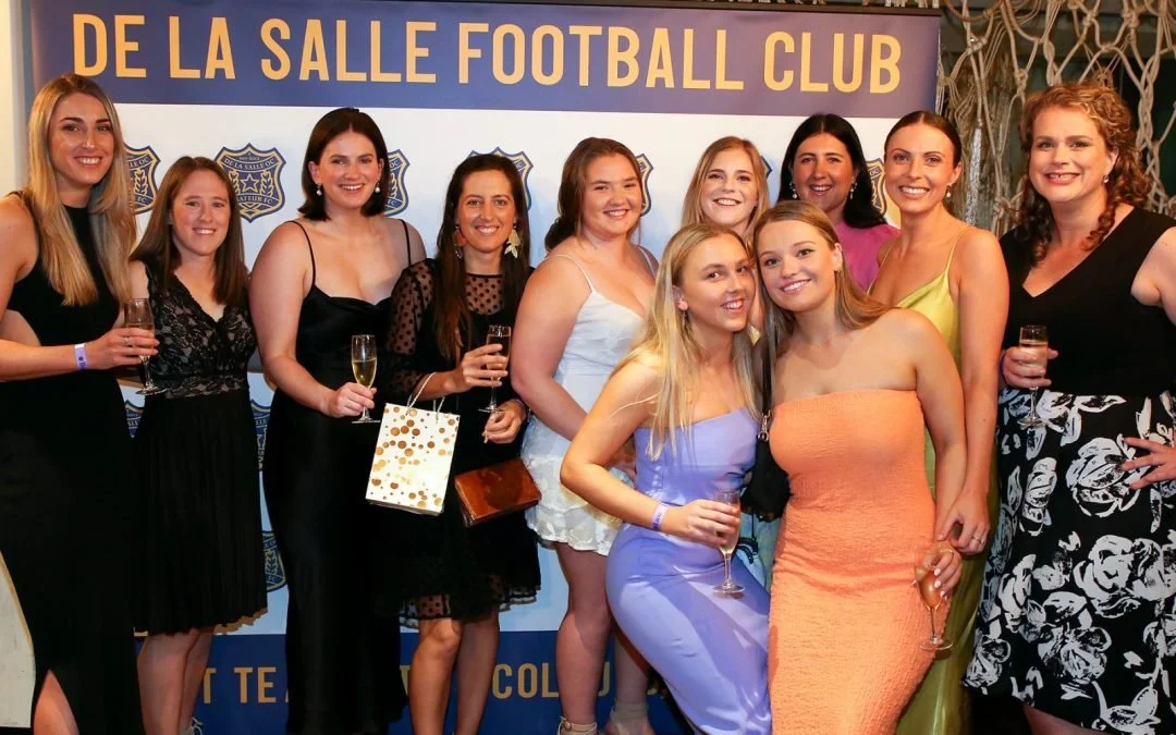 Group of women at a celebration event for De La Salle Football Club, posing in front of a banner, holding glasses of champagne and smiling.